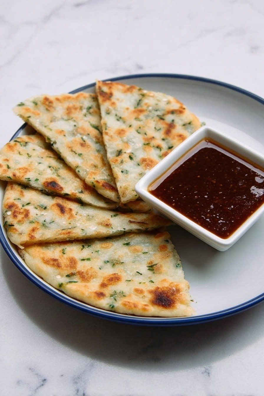 The image shows four pieces of flatbread-like food arranged in a fan shape on a white plate with a blue inside. The flatbreads are light golden-brown with some green herbs visible in the dough, showing a soft and slightly bumpy texture. On the plate, near the upper-right side of the flatbreads, there is a small white square bowl filled with a dark brown sauce that looks smooth and glossy. The plate is placed on a white marbled surface, creating a clean and simple background. photo taken with an iphone --ar 2:3 --v 7 - Chinese Scallion Pancakes, How to make Chinese scallion pancakes, crispy scallion pancakes recipe, homemade scallion pancakes, Chinese flatbread recipes