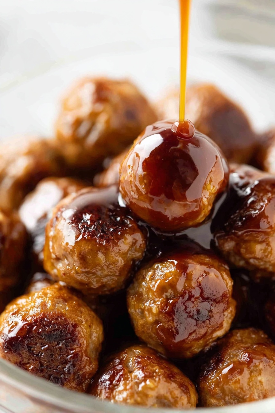 A close-up view of a pile of shiny brown meatballs in a clear bowl on a white marbled surface, with a thick reddish-brown sauce being poured slowly over the top meatball in the center. The meatballs are round and vary slightly in size, with a glazed texture showing small char marks and a glossy finish from the sauce. The sauce is thick and smooth, creating a shiny coating that drips down the sides of the meatballs. The image focuses closely on the meatballs with a soft blurry background. Photo taken with an iphone --ar 2:3 --v 7 - Sweet and Sour Cocktail Meatballs, cocktail meatballs recipe, easy appetizer recipes, party meatball ideas, flavorful meatball recipes