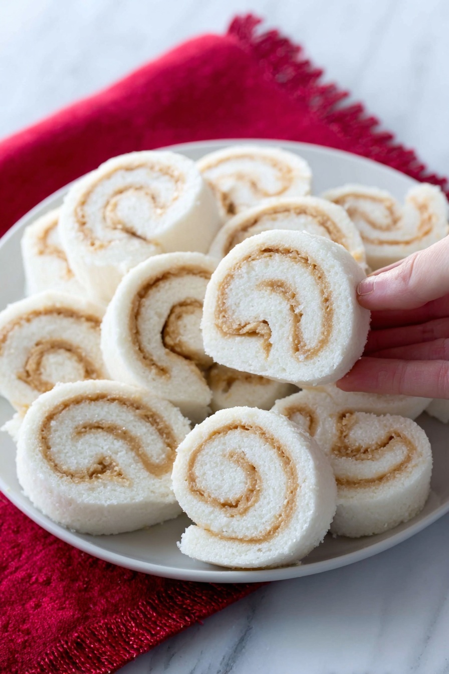 The image shows a white plate filled with soft, spiral-shaped rolls arranged closely together. Each roll has a smooth white outer layer with swirled light tan filling inside, creating a clear contrast. The rolls look fluffy and neatly sliced, with the spirals visible on the sides. The plate is placed on a bright red cloth, and one roll is in the foreground, slightly separated. The background and surface have a white marbled texture. A woman's hand is holding one roll in the top right corner. photo taken with an iphone --ar 2:3 --v 7 - Potato Candy Pinwheels with Peanut Butter, nostalgic candy recipes, easy potato candy, homemade peanut butter pinwheels, vintage dessert ideas