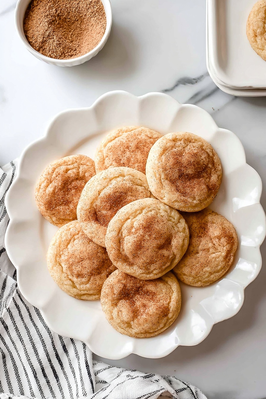 There are seven round snickerdoodle cookies with a light golden brown color and a dusting of cinnamon sugar on top, arranged in a flower shape on a white scalloped plate. The cookies have a soft, slightly wrinkled texture and are placed on a white marbled surface. Near the top left of the plate, there is a small white bowl filled with cinnamon sugar. A white cloth with thin black stripes lays slightly under the plate on the left side. Photo taken with an iphone --ar 2:3 --v 7 - Chewy Snickerdoodle Cookies, Snickerdoodle Cookies recipe, cinnamon sugar cookies, soft chewy cookies, easy cookie recipes