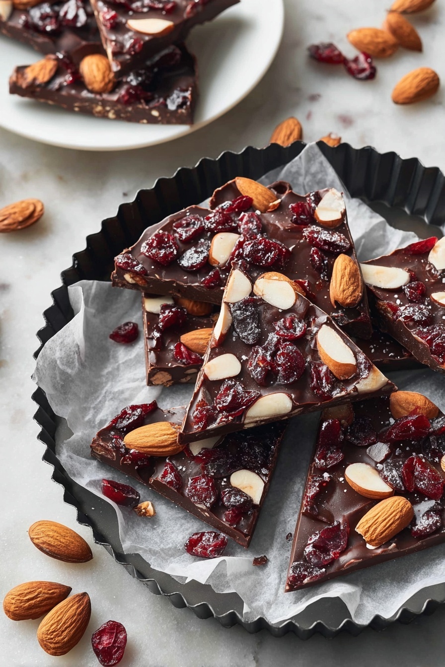 The image shows several pieces of chocolate bark, each with one thick layer of dark brown chocolate topped with whole light brown almonds and dark red dried cranberries scattered evenly. The chocolate pieces are irregular triangles and squares placed on white parchment paper inside a black fluted round pan. Around the pan are whole almonds and dried cranberries on a white marbled textured surface. In the background, a white plate holds three more pieces of chocolate bark, showing the same dark chocolate base with almonds and cranberries. photo taken with an iphone --ar 2:3 --v 7 - Easy Chocolate Bark with Nuts and Dried Fruit, chocolate bark with nuts and dried fruit, homemade chocolate bark, quick chocolate snack, holiday chocolate bark