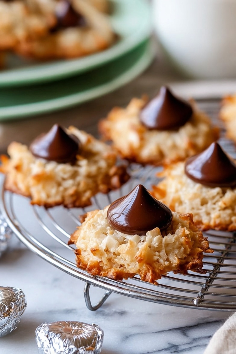 The image shows several round cookies with a rough, golden-brown texture from toasted coconut flakes, each with a smooth, shiny dark chocolate drop placed in the center. The cookies sit on a silver wire cooling rack, which rests on a white marbled surface. Around the rack, there are small foil-wrapped chocolate drops scattered. The background includes a blurred white bowl and a soft green plate with more cookies, creating a warm and inviting scene. photo taken with an iphone --ar 2:3 --v 7 - Coconut Macaroon Blossoms with Hershey Kisses, coconut macaroon cookies, chocolate surprise cookies, easy gluten-free cookies, holiday cookie recipes