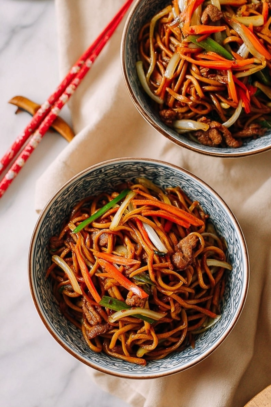Two white bowls with blue patterns on the edges hold a serving of stir-fried noodles. The dish has multiple layers: a base of dark brown soy-coated noodles entwined with thin strips of orange carrots, white onions, and light green vegetables. Small pieces of cooked brown meat are scattered throughout. Both bowls are placed on a soft beige cloth on a white marbled surface. A pair of red and cream chopsticks rest near the bowls. photo taken with an iphone --ar 2:3 --v 7 - Restaurant-Style Chicken Lo Mein, chicken lo mein recipe, Chinese chicken lo mein, easy lo mein stir-fry, homemade Chinese noodles