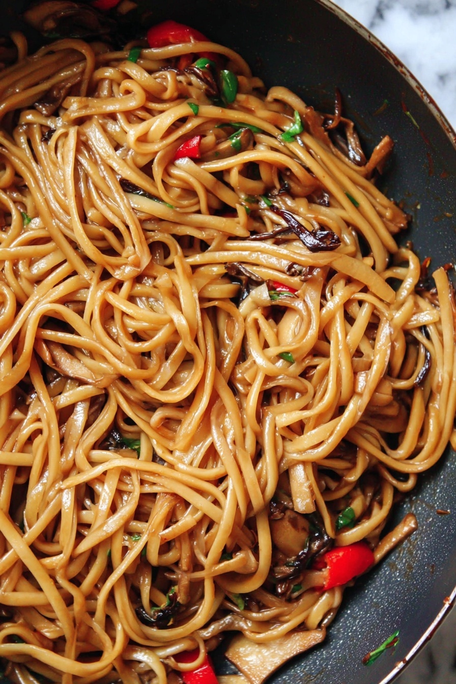 The image shows a close-up of a pan filled with long, shiny noodles coated in a light brown sauce. Mixed among the noodles are small pieces of light tan tofu, dark brown thin mushroom strips, and bright red chili slices, with tiny bits of green herbs scattered throughout. The noodles look soft and glossy, with a slight twist and curl to them, giving a textured, tangled appearance. The pan has a dark surface and the background is changed to a white marbled texture. photo taken with an iphone --ar 2:3 --v 7 - Vegan Kung Pao Pasta with Mushrooms, vegan pasta recipes, spicy mushroom pasta, plant-based Kung Pao sauce, quick vegan dinner ideas