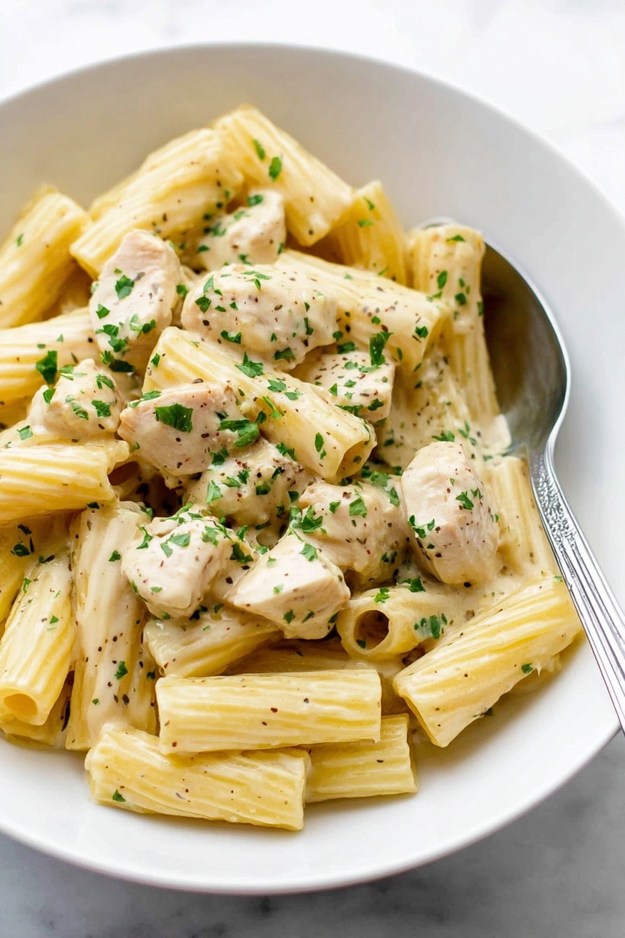 A white bowl filled with creamy rigatoni pasta mixed with chunks of light beige chicken pieces, all coated in a smooth, pale yellow sauce. The pasta is ribbed and tubular, scattered evenly throughout the dish. Small green parsley flakes are sprinkled over the top, adding bright color contrast. A silver spoon rests inside the bowl on the right side, partially under the pasta. The bowl sits on a white marbled surface. Photo taken with an iphone --ar 2:3 --v 7 - Creamy Lemon Chicken Pasta, Lemon Chicken Pasta, Easy Pasta Dinner, Quick Chicken Pasta, Zesty Creamy Pasta
