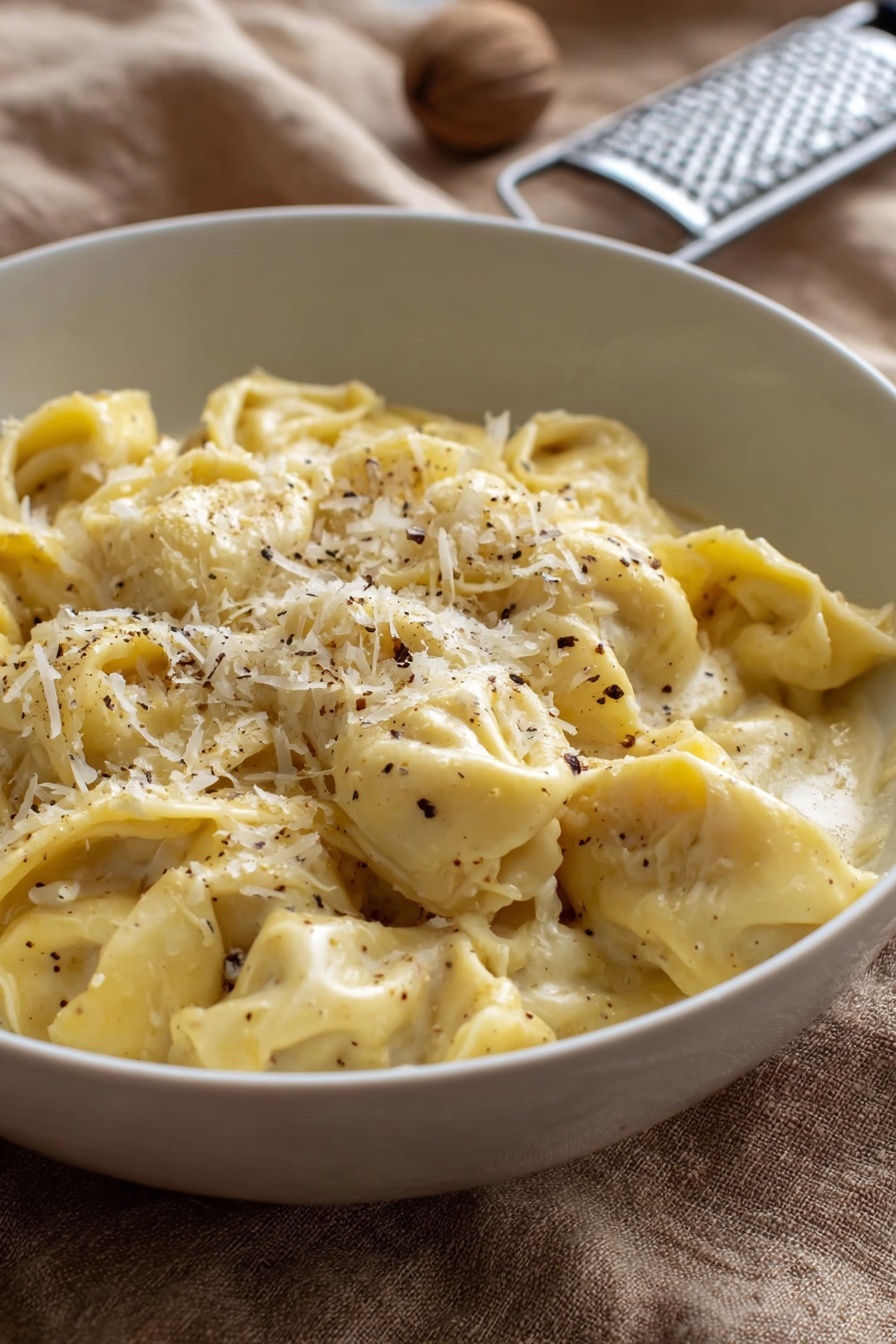 A white bowl filled with about two layers of soft, folded tortellini pasta in a creamy, pale yellow sauce. The pasta is sprinkled with finely grated white cheese and small black pepper flakes, mostly concentrated on the top center. The bowl rests on a tan textured cloth, with a metal grater and a whole nutmeg blurred in the background. The lighting is natural and soft, showing the smooth creamy texture of the sauce and the delicate folds of the tortellini photo taken with an iphone --ar 2:3 --v 7 - Creamy Cheese Tortellini, cheese tortellini recipe, easy creamy pasta, quick comfort food, cheesy pasta dish