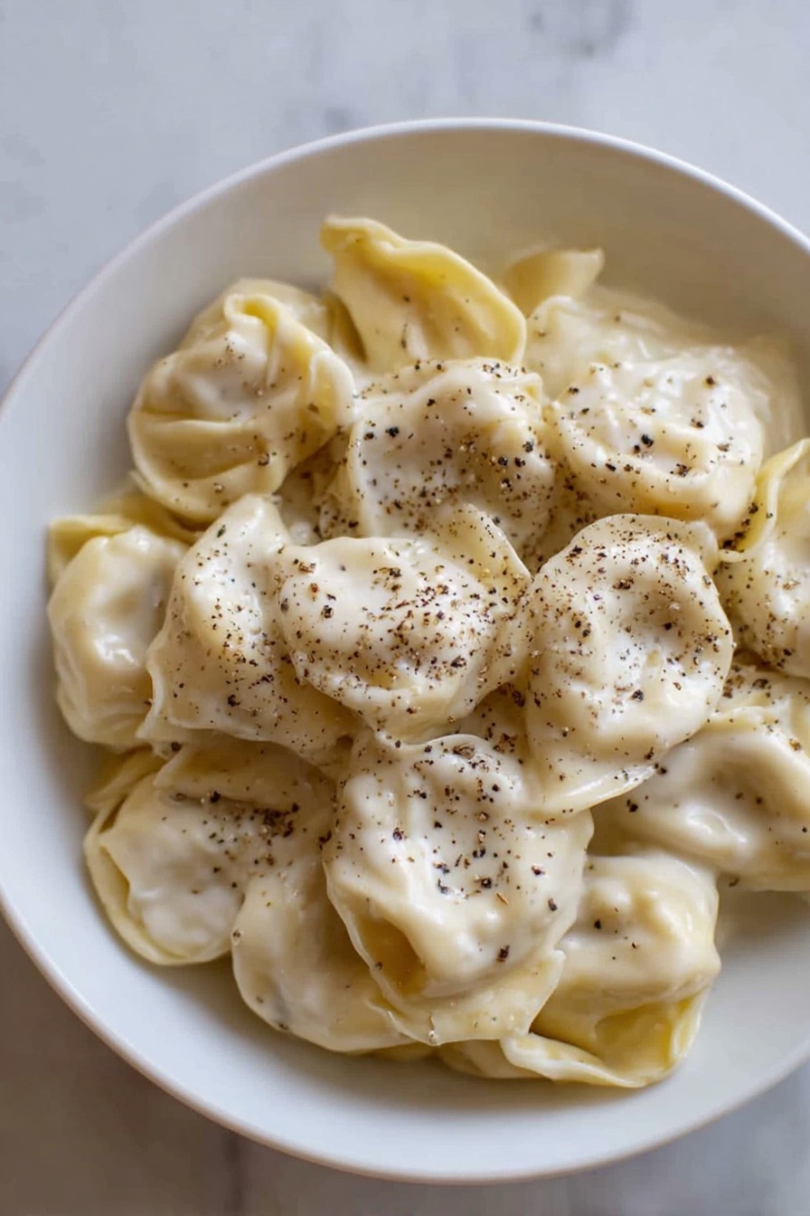 A white bowl filled with a single layer of tortellini pasta, covered evenly with a smooth, creamy white sauce that has a slightly shiny texture. The tortellini are pale yellow with soft folds, and black pepper is sprinkled generously on top, adding small dark specks scattered over the sauce. The bowl is placed on a white marbled surface, and the light highlights the soft texture of both the pasta and sauce photo taken with an iphone --ar 2:3 --v 7 - Creamy Cheese Tortellini, cheese tortellini recipe, easy creamy pasta, quick comfort food, cheesy pasta dish