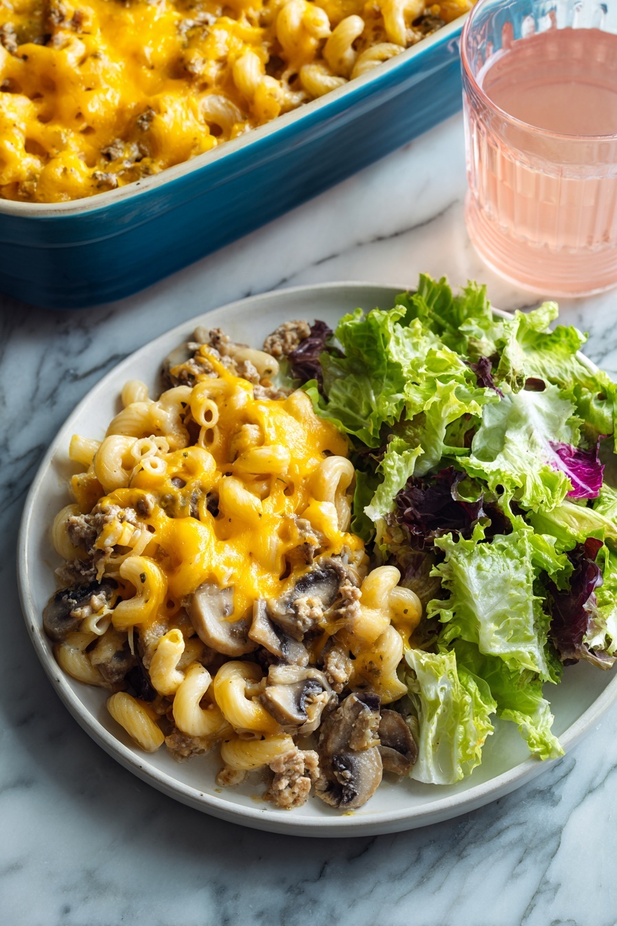 The image shows a white round plate with two main parts: on the left, a layer of baked macaroni mixed with ground meat and sliced mushrooms, all topped with melted bright yellow-orange cheese that looks gooey and soft, and on the right, a fresh green salad made of different lettuce leaves with a mix of light and dark green colors and some purple edges. The plate is set on a white marbled surface, and in the background, there is a blue baking dish filled with more of the macaroni bake and a clear glass of light pink drink. Photo taken with an iphone --ar 2:3 --v 7 - Beef Mushroom Johnny Marzetti Casserole, hearty beef mushroom pasta bake, easy comfort food casserole, cheesy ground beef and mushroom casserole, family favorite dinner