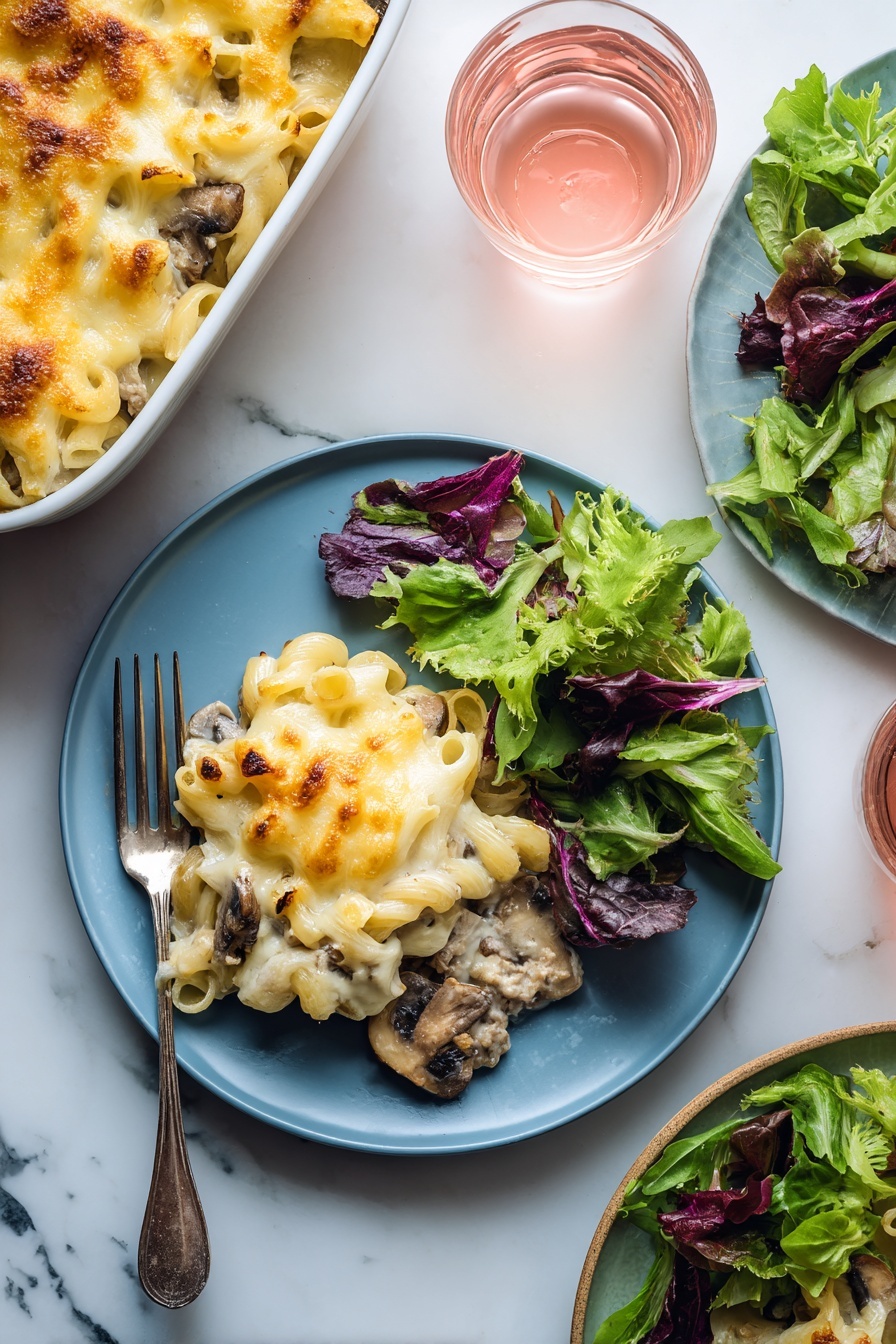 A blue plate holds a serving of baked macaroni and cheese with visible pieces of mushrooms and ground meat under a layer of melted golden-yellow cheese on the left side; on the right side of the plate, there is a fresh green salad made of mixed leafy greens with different shades of green and purple. A silver fork rests on the plate's left edge underneath the macaroni. In the background, a white baking dish filled with more macaroni and cheese sits on a white marbled surface along with a small clear glass of light pink drink. Another plate with leafy greens and another glass of pink drink are partially visible on the right side. photo taken with an iphone --ar 2:3 --v 7 - Beef Mushroom Johnny Marzetti Casserole, hearty beef mushroom pasta bake, easy comfort food casserole, cheesy ground beef and mushroom casserole, family favorite dinner
