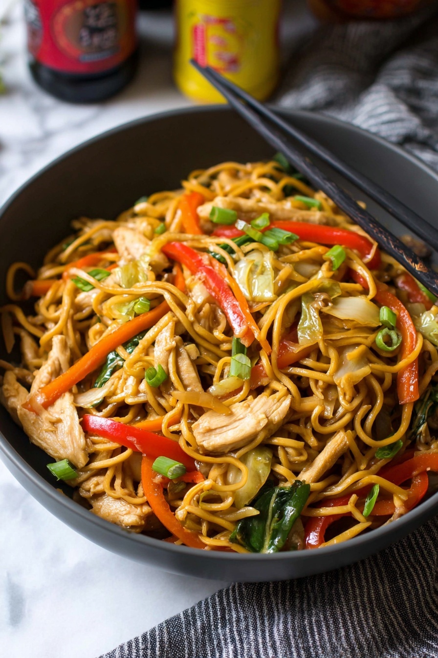 A close-up view of a dark gray bowl filled with stir-fried noodles mixed with thin strips of light beige chicken pieces, bright red bell pepper slices, green scallion pieces, thin orange carrot strips, light green cabbage, and sliced beige mushrooms. Black chopsticks rest on the right edge of the bowl, while the bowl sits on a white marbled surface with part of a gray striped cloth to the right. The background includes some blurred bottles with yellow and red labels. photo taken with an iphone --ar 2:3 --v 7 - Chicken Yakisoba Stir-Fry, Japanese stir-fry recipes, easy chicken noodle stir-fry, quick Asian dinner ideas, simple yakisoba with chicken