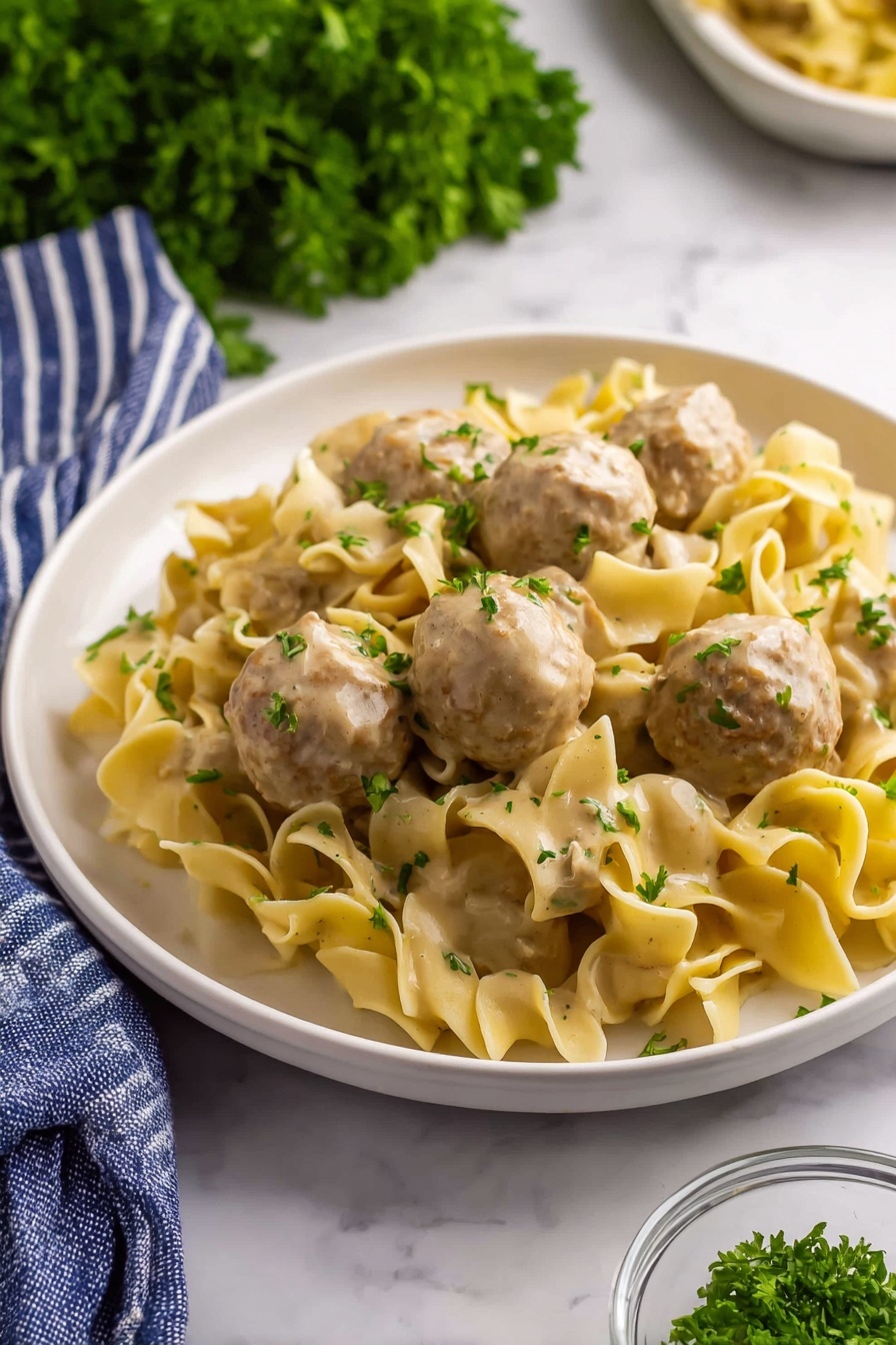 A white plate holds a serving of wide, curly egg noodles mixed with round meatballs covered in a creamy light brown sauce. The noodles are soft and slightly twisted, with flecks of green herbs sprinkled on top. The meatballs are light brown, smooth, and sit evenly spaced among the noodles. Bright green parsley leaves are scattered across the dish, adding color contrast. The scene is set on a white marbled surface with a bunch of fresh parsley blurred in the background, a blue and white striped cloth partially visible at the bottom left, and a small glass bowl with chopped herbs near the plate. Photo taken with an iphone --ar 2:3 --v 7 - Creamy Swedish Meatball Noodle Skillet, Swedish meatball skillet, creamy meatball dish, one-pan Swedish meatballs, comfort food recipes