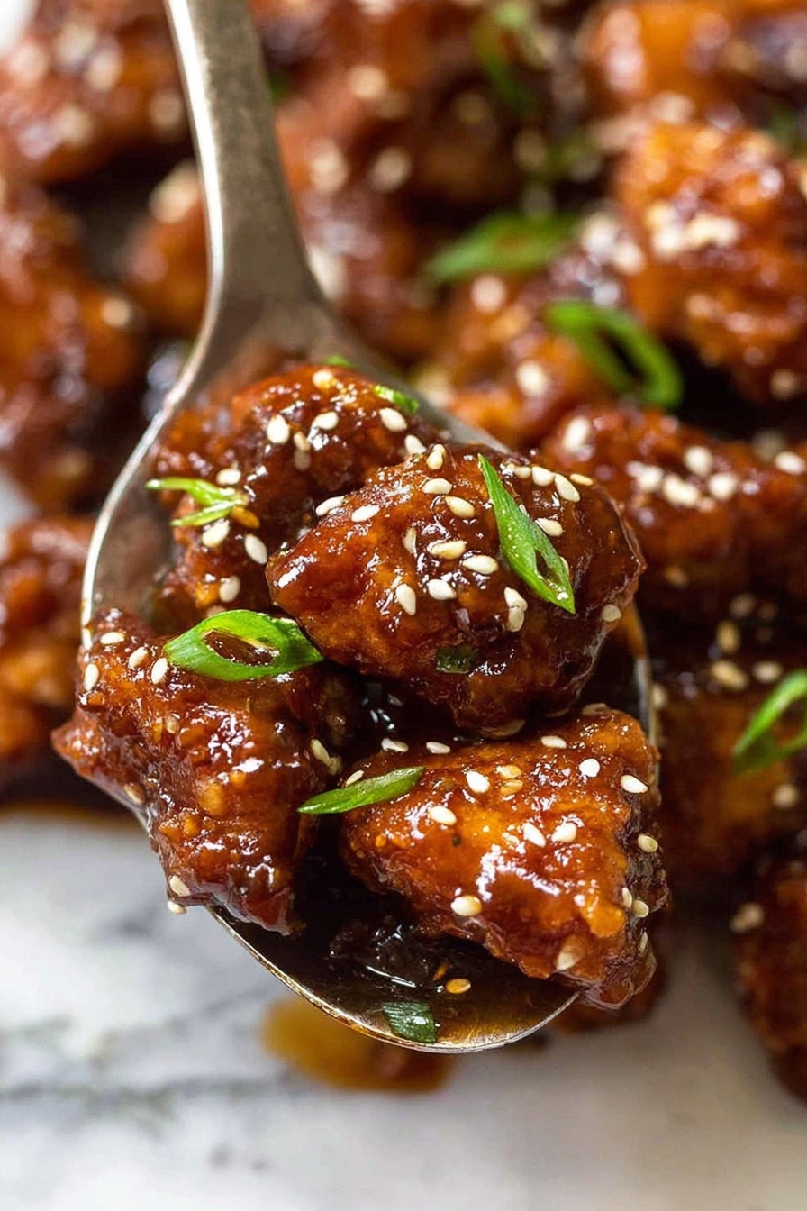 The image shows a close-up view of small pieces of fried chicken coated in a thick, shiny, dark brown sauce. The chicken pieces are irregular in shape with a crispy texture underneath the sauce. White sesame seeds are scattered on top, adding contrast and texture. Thin green slices of scallions or green onions are sprinkled over the chicken, providing a splash of fresh color. The chicken rests in a metal spoon, with more saucy chicken pieces visible in the background, all set on a white marbled surface. photo taken with an iphone --ar 2:3 --v 7 - Crispy General Tso's Chicken, homemade General Tso's chicken, best crispy chicken recipes, spicy sweet chicken dish, Asian chicken stir-fry