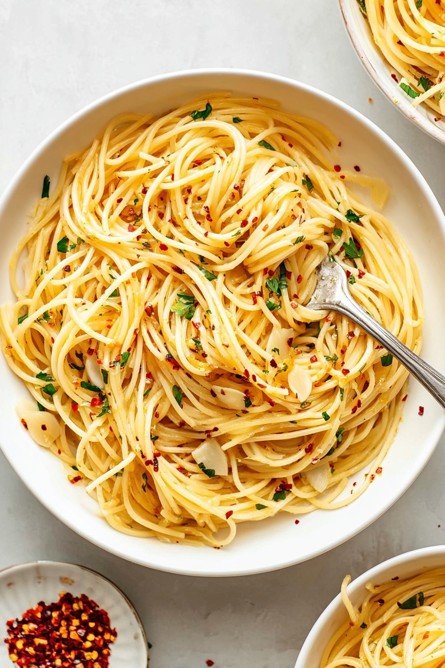 A large white bowl filled with long, thin spaghetti noodles that are lightly coated with olive oil, giving them a soft yellow color. Scattered throughout the pasta are thin, flat slices of garlic and small bits of fresh green parsley. Tiny red chili flakes are sprinkled evenly on top, adding small pops of red. A silver fork is twirling a small bundle of the pasta near the center of the bowl. The bowl rests on a white marbled surface, with a small white plate next to it containing additional red chili flakes. Part of another similar bowl filled with the same pasta can be seen in the upper right corner. Photo taken with an iphone --ar 2:3 --v 7 - Garlic Spaghetti Aglio e Olio, easy garlic pasta, quick spaghetti recipes, simple Italian pasta, flavorful garlic spaghetti
