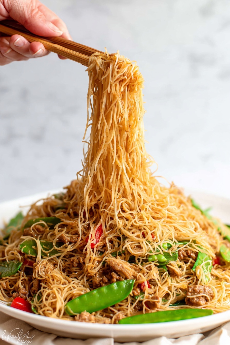 The image shows a white plate full of thin noodles mixed with small pieces of cooked meat, green snow peas, and red bell pepper slices. The noodles are light brown and soft, forming the top and main layer, while the vegetables and meat are scattered throughout, adding pops of green and red colors. A woman's hand is holding wooden chopsticks lifting a portion of the noodles above the plate, with the noodles hanging down in thin strands. The background has a white marbled texture. photo taken with an iphone --ar 2:3 --v 7 - Singapore Curry Noodles with Chicken, Singapore Curry Noodles, Curry Noodles with Chicken, Asian Curry Noodles, Quick Curry Noodle Recipe