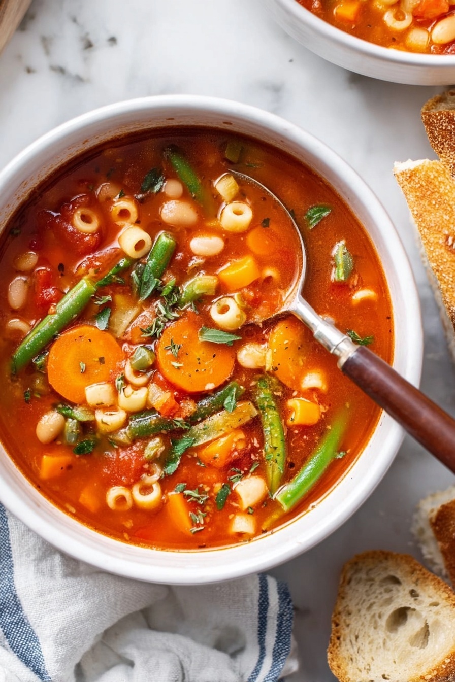A white bowl filled with a bright red tomato-based soup with visible small round pasta pieces, sliced orange carrots, white beans, green beans, and finely chopped herbs. A silver spoon with a brown handle is placed inside the bowl, resting on the side. The bowl sits on a white marbled surface. To the right, rustic pieces of sliced bread are partially visible, and on the left, a soft white cloth with blue stripes is partly in the frame. photo taken with an iphone --ar 2:3 --v 7 - Hearty Vegetable Minestrone Soup, vegetable minestrone recipe, healthy vegetable soup, hearty veggie soup, comforting minestrone