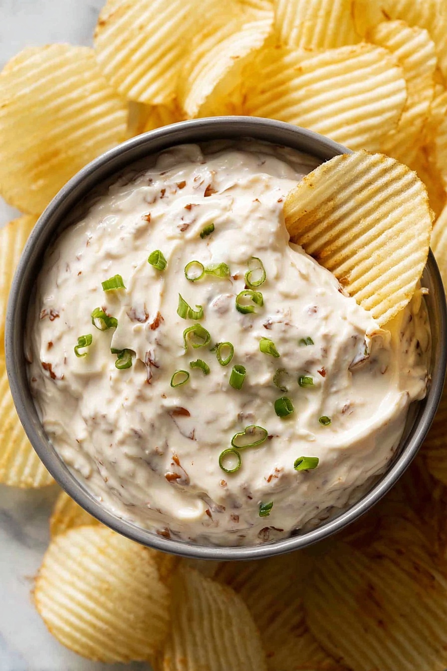A gray round bowl filled with creamy dip that has visible small brown bits throughout, topped with finely chopped green onions scattered on the surface. A white ridged potato chip is dipped into the creamy mixture on the right side of the bowl. The bowl is surrounded by more white ridged potato chips, all arranged on a white marbled surface. photo taken with an iphone --ar 2:3 --v 7 - Homemade French Onion Dip, easy French onion dip, creamy onion dip recipe, homemade party dip, savory onion dip for snacks