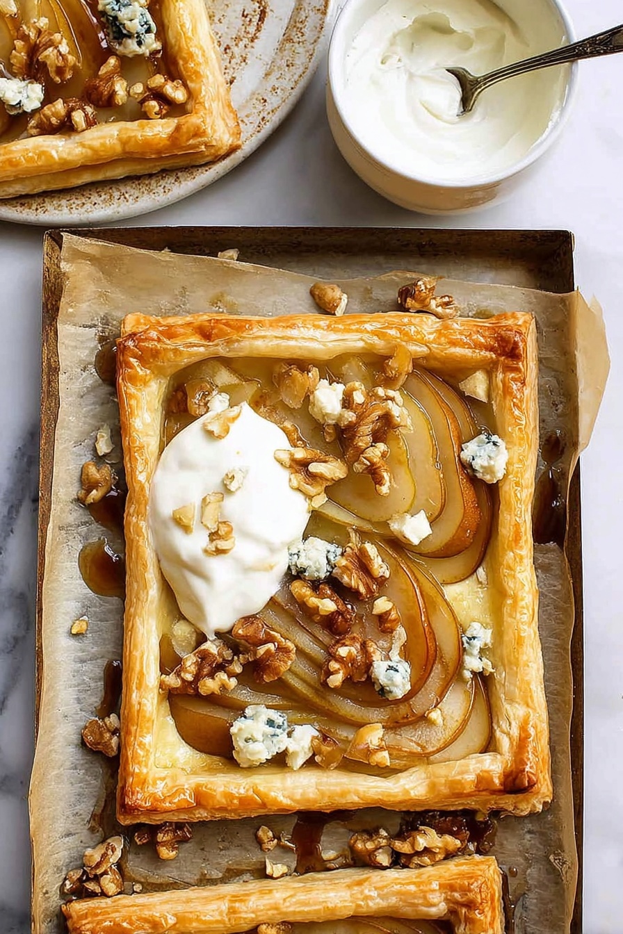 The image shows a golden brown puff pastry tart with a raised edge surrounding soft, thinly sliced caramelized pears arranged in an overlapping pattern. On top of the pears, there are small chunks of blue cheese and scattered walnut pieces adding texture and color contrast. A dollop of creamy white yogurt or cream is placed on one corner of the tart. The tart sits on parchment paper which lines a baking tray with some walnut pieces and cheese crumbs around it. Nearby, a small white ceramic bowl with a spoon inside contains more white cream. The background is a white marbled surface. photo taken with an iphone --ar 2:3 --v 7 - Caramelized Pears and Blue Cheese Tart, pear and blue cheese tart, caramelized pear dessert, fancy tart recipes, elegant fruit tart