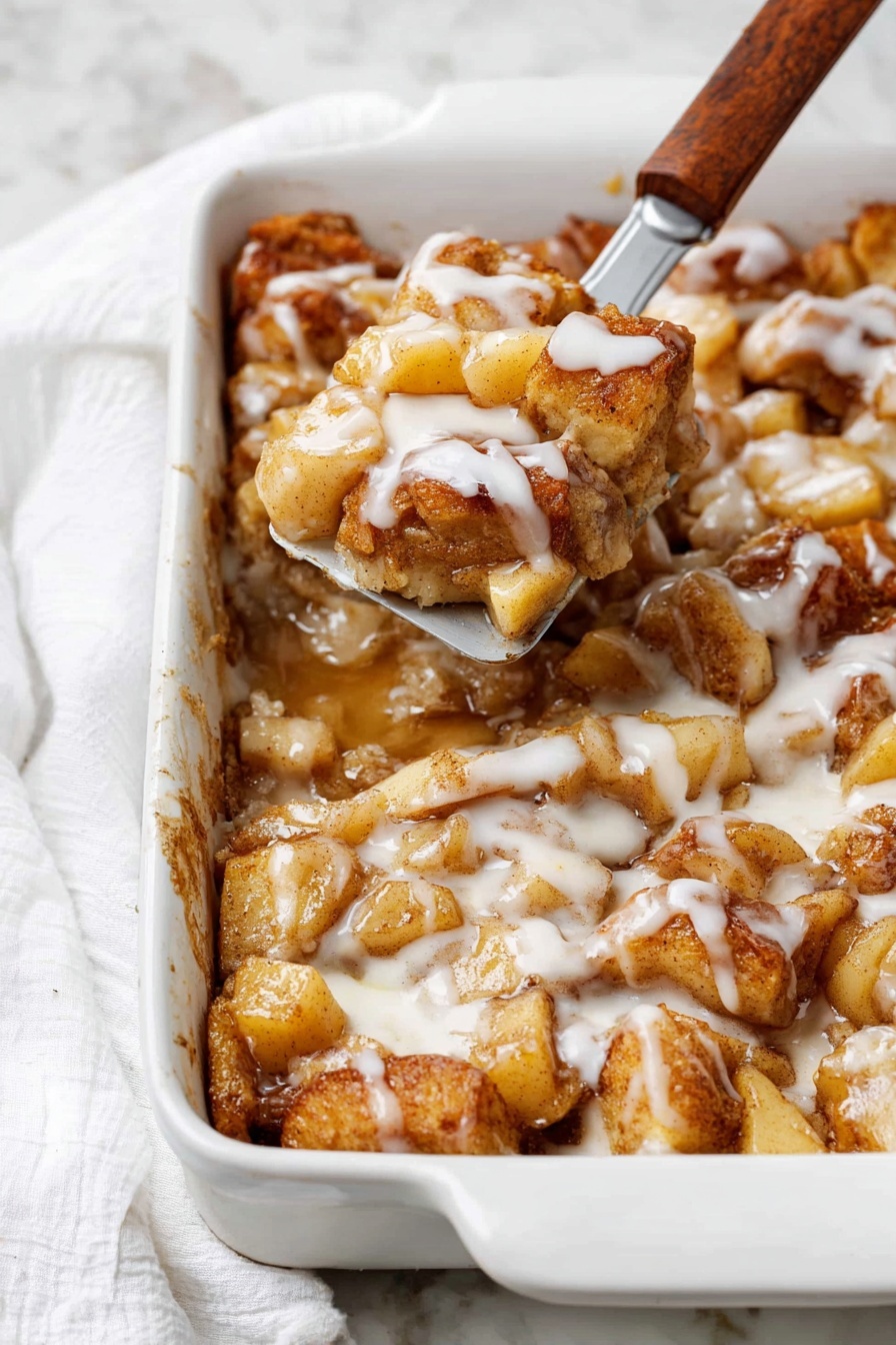A white rectangular baking dish filled with a baked dessert made of soft, golden brown pieces layered with light brown cooked apple chunks and drizzled generously with a white creamy glaze. The dessert has a slightly sticky texture from syrup or sauce that pools around the pieces. A metal spatula with a wooden handle lifts a portion showing the soft inside layers and more apples. The background is a white marbled surface with a white cloth nearby. photo taken with an iphone --ar 2:3 --v 7 - Apple Pie Cinnamon Roll Bake, fall breakfast recipes, easy cinnamon roll bake, apple dessert casseroles, cozy brunch ideas
