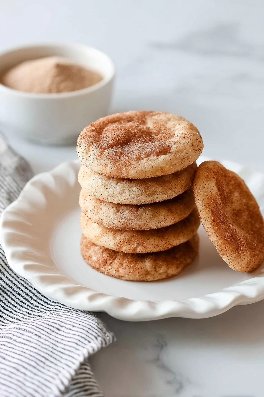 A stack of eight round, light brown cookies with a dusting of cinnamon sugar on top sits in the center of a white plate with scalloped edges. One cookie lies flat behind the stack, showing its darker cinnamon spots. The white plate is placed on a white marbled surface with a folded white and black striped cloth partially visible on the left. In the top left corner, a small white bowl filled with a light brown powder is slightly out of focus. photo taken with an iphone --ar 2:3 --v 7 - Chewy Snickerdoodle Cookies, Snickerdoodle Cookies recipe, cinnamon sugar cookies, soft chewy cookies, easy cookie recipes