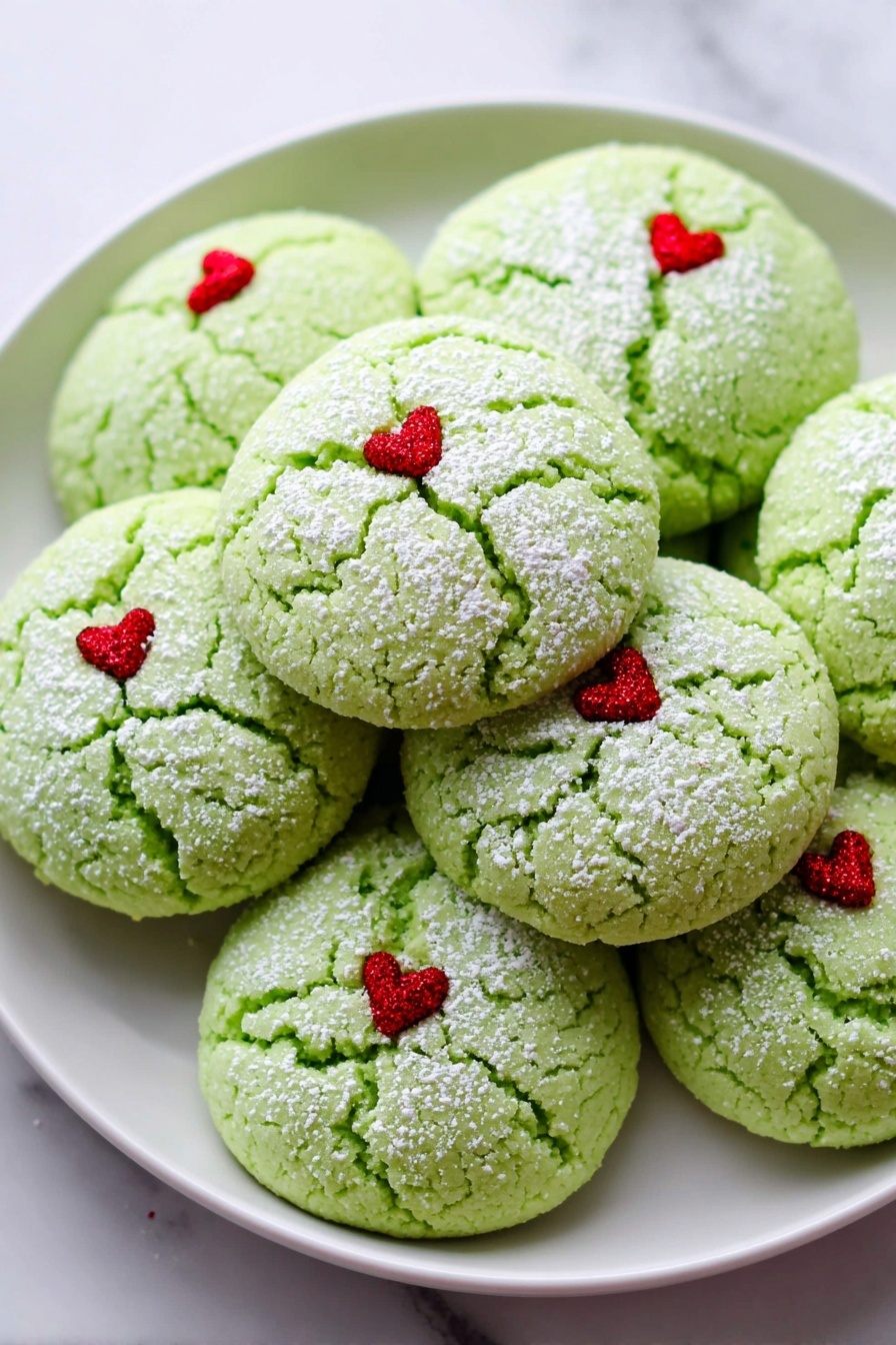 The image shows a white plate filled with round green cookies that have a cracked texture on top. Each cookie has a light dusting of white powder and a tiny red heart decoration placed slightly off-center on the top surface. The cookies are stacked closely together on the plate, which sits on a white marbled background. Photo taken with an iphone --ar 2:3 --v 7 - Grinch Cookies with Mint and Heart Sprinkles, holiday cookies with mint and heart decorations, festive green Christmas cookies, easy holiday cookie recipes, minty holiday treats