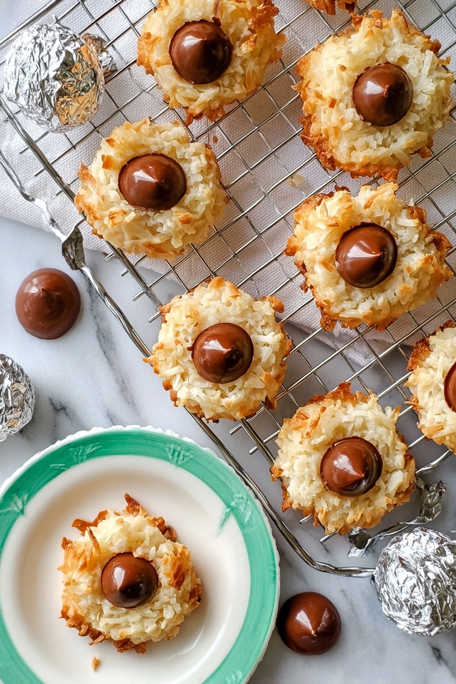 The image shows seven round coconut cookies with a light golden-brown crispy outside and white, shredded coconut texture in the middle layer. Each cookie has a single smooth milk chocolate drop placed in the center on top. The cookies are arranged on a silver cooling rack that rests on a white rectangular marble slab. Around the rack, there are several extra chocolate drops scattered on the white marbled surface. A woman's hand is holding one cookie on a white plate with a green rim, and a few silver-wrapped chocolate candies are placed nearby. The overall setting is bright with a white marbled background. photo taken with an iphone --ar 2:3 --v 7 - Coconut Macaroon Blossoms with Hershey Kisses, coconut macaroon cookies, chocolate surprise cookies, easy gluten-free cookies, holiday cookie recipes