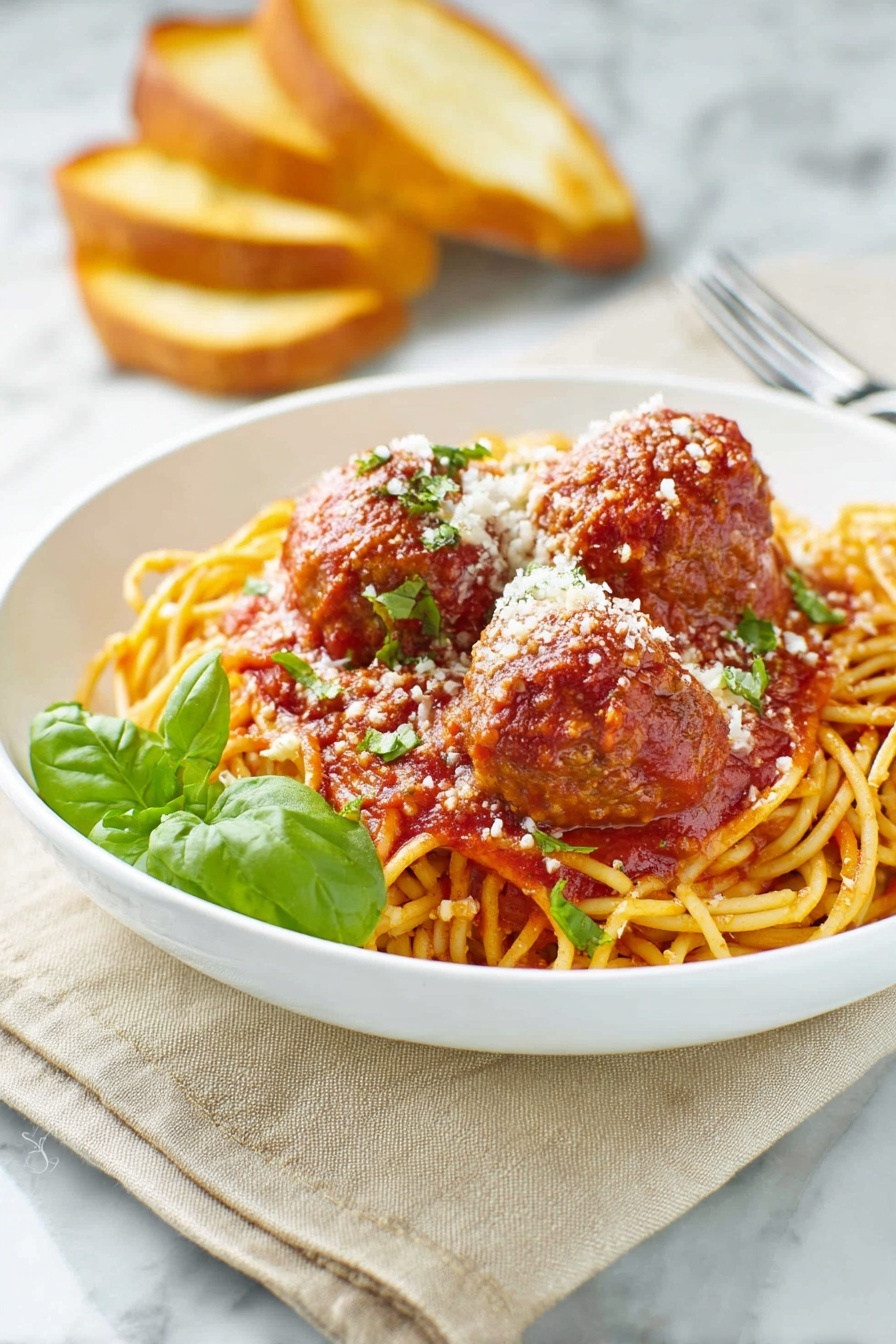 A white bowl holds a bed of golden-yellow spaghetti noodles topped with rich red tomato sauce. On top, there are three large brown meatballs coated in the sauce, sprinkled with white grated cheese and small green herb pieces. A fresh green basil leaf adds color near the front. In the background, there are three toasted bread slices leaning against the bowl. The scene sits on a beige cloth over a white marbled surface. A silver fork rests nearby on the right side. Photo taken with an iphone --ar 2:3 --v 7 - Homemade Spaghetti and Meatballs, Italian spaghetti and meatballs, classic spaghetti with meatballs, homemade marinara sauce, easy Italian dinner