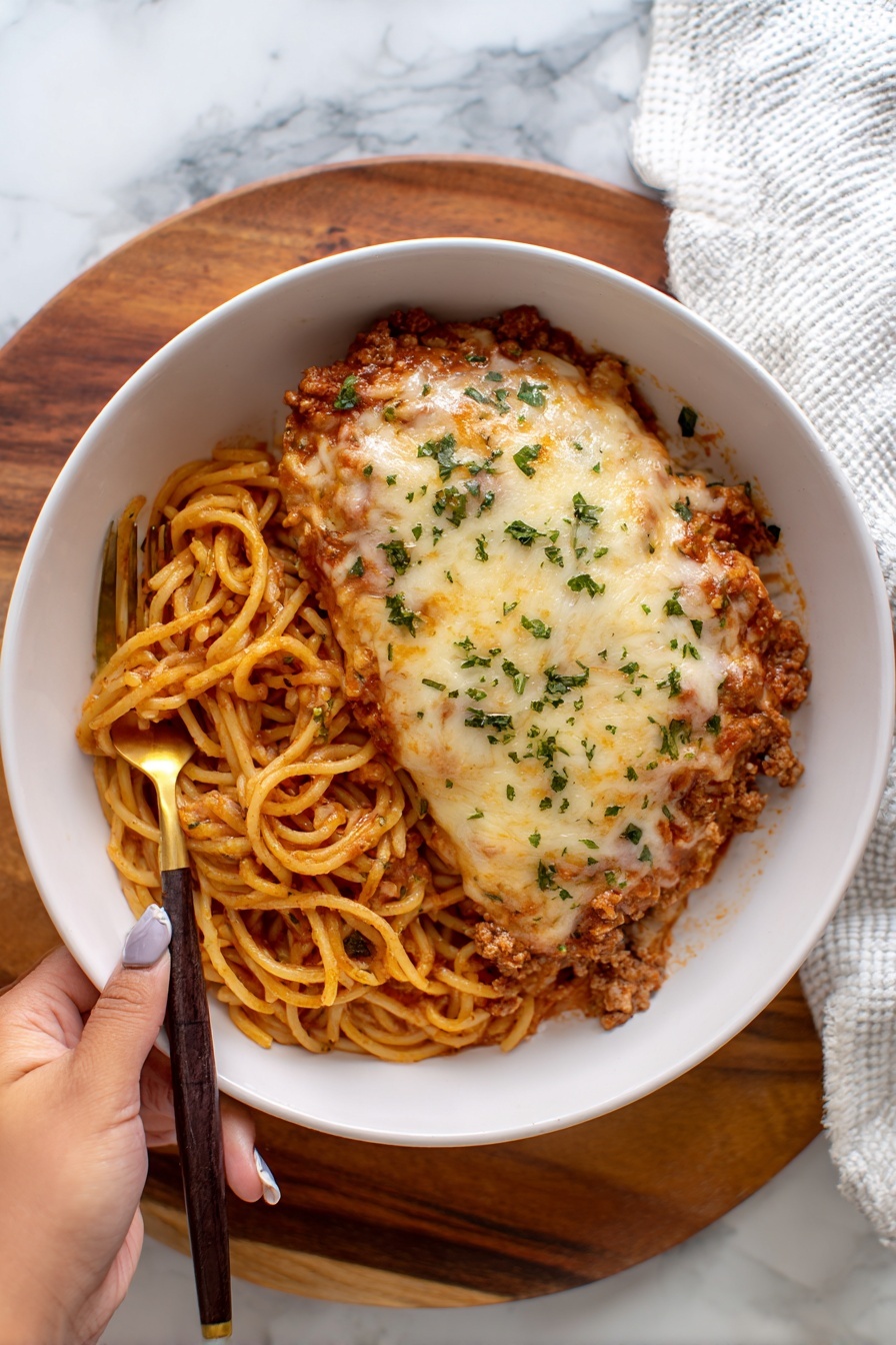 A white rectangular baking dish filled with a layered lasagna, showing golden-brown melted cheese on top with some darker browned spots and red tomato sauce peeking through. The edges are bubbly and slightly crispy, showing a textured mix of cheese and sauce. Around the dish, there is a beige cloth beneath, and on the white marbled surface near the top-left corner, there are two small white bowls, one filled with fresh green parsley and the other with thin white slices, possibly cheese. photo taken with an iphone --ar 2:3 --v 7 - Cheesy Loaded Baked Spaghetti, baked spaghetti casserole, cheesy pasta bake, easy baked spaghetti recipe, hearty spaghetti dinner