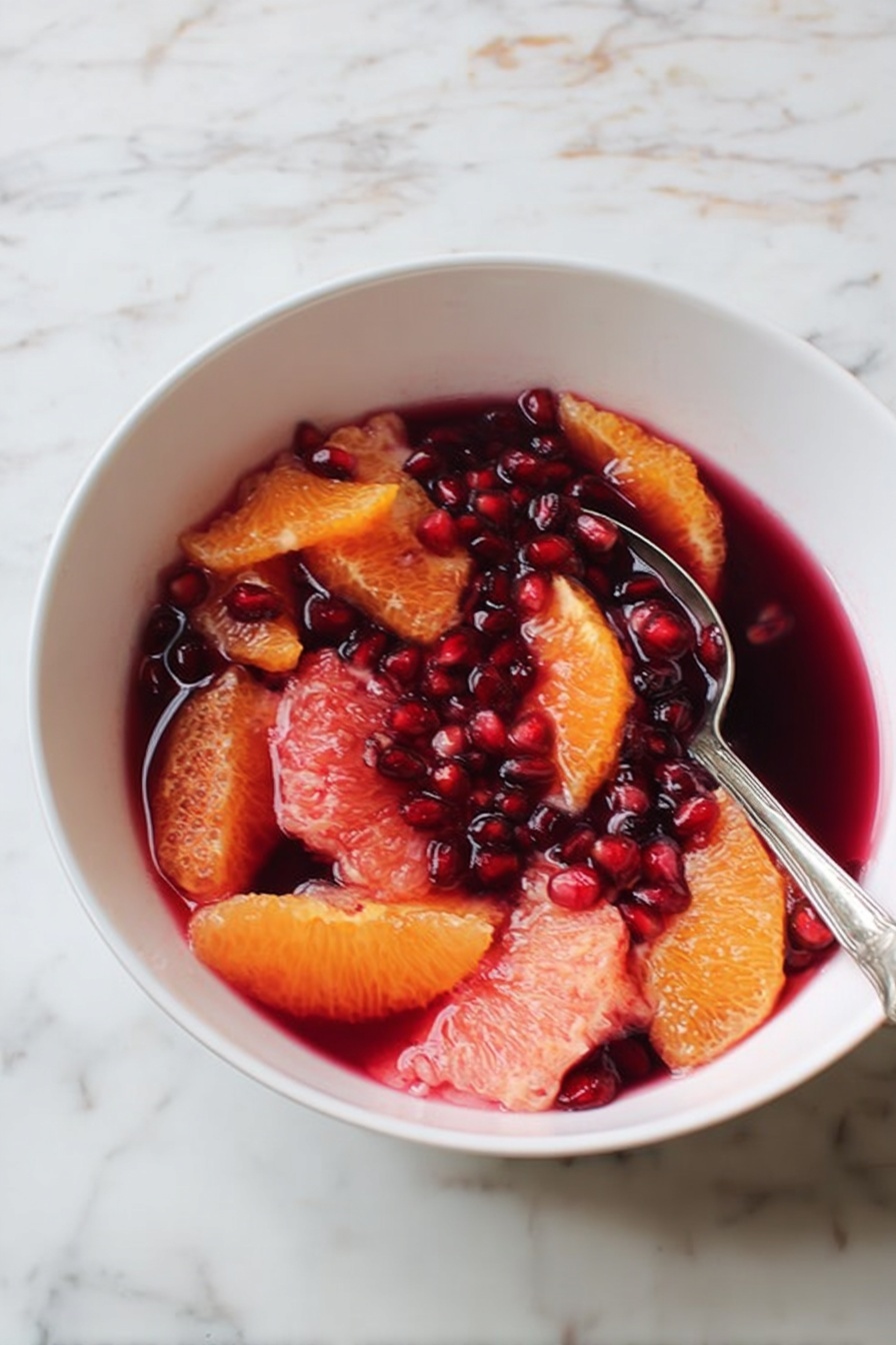 A white bowl filled with two main layers: the bottom layer is a deep red liquid, and floating on top are bright orange and pink citrus slices along with many shiny dark red pomegranate seeds. A silver spoon rests inside the bowl on the right side. The bowl is placed on a white marbled surface with soft veins of gray and faint brown. photo taken with an iphone --ar 2:3 --v 7 - Winter Citrus & Pomegranate Salad, citrus pomegranate salad, winter fruit salad, healthy citrus pomegranate salad, colorful winter salad