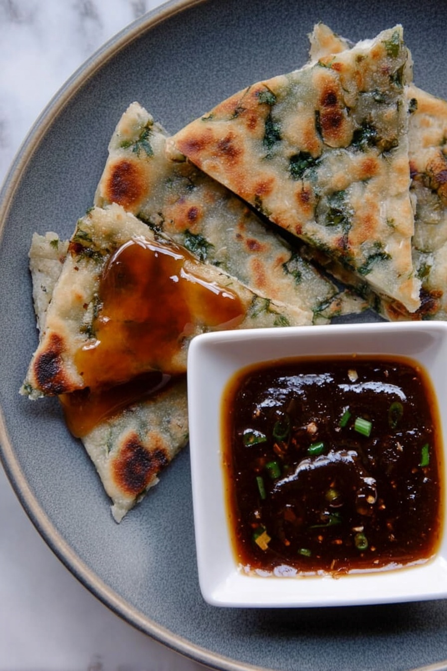 The image shows a close-up of a grey plate on a white marbled surface, with four triangular pieces of flatbread that have green herbs inside, each piece varying in size and texture with some browned spots from cooking. One piece near the bottom left has a shiny layer of sticky, dark amber sauce on top. To the right, a small white square bowl holds a thick, dark brown dipping sauce with small green bits inside, creating a glossy surface. photo taken with an iphone --ar 2:3 --v 7 - Chinese Scallion Pancakes, How to make Chinese scallion pancakes, crispy scallion pancakes recipe, homemade scallion pancakes, Chinese flatbread recipes