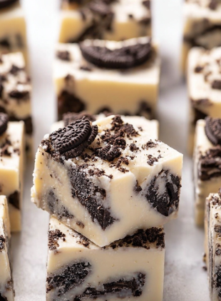 The image shows a close-up of thick square pieces of cookies and cream fudge layered in white creamy base mixed with broken chocolate cookie bits. Each piece has a smooth, pale off-white layer filled with dark brown cookie chunks inside and baked bits scattered on top, creating a textured look with irregular shapes. The fudge pieces are placed closely together on a white marbled surface, highlighting the contrast between the creamy white fudge and dark cookies. Photo taken with an iphone --ar 2:3 --v 7 - White Chocolate Oreo Fudge, Oreo fudge recipe, easy Oreo fudge, no-bake Oreo fudge, creamy Oreo fudge