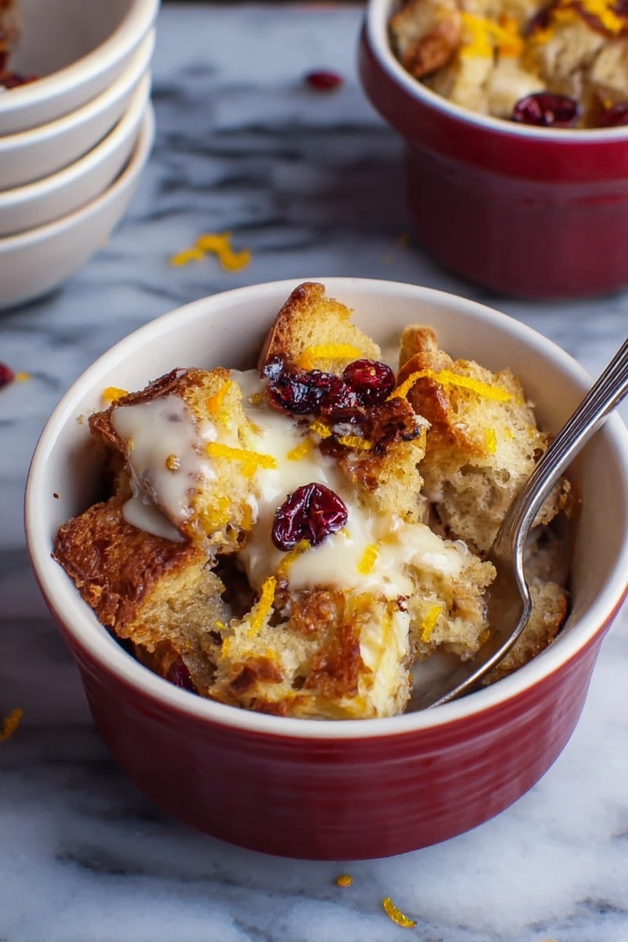 A small white bowl filled with several layers of toasted bread chunks that are golden brown and crispy on the edges. The bread pieces are mixed with creamy, slightly melted white sauce or cream that sits on top and around the bread. There are dark red dried berries scattered on top and some thin orange zest strips add a pop of color. A silver spoon is inserted inside the bowl on the right side. The bowl is placed on a white marbled surface with a small stack of empty white bowls in the background. photo taken with an iphone --ar 2:3 --v 7 - Cranberry Bread Pudding, holiday dessert recipes, easy bread pudding, festive cranberry dessert, cozy holiday treats