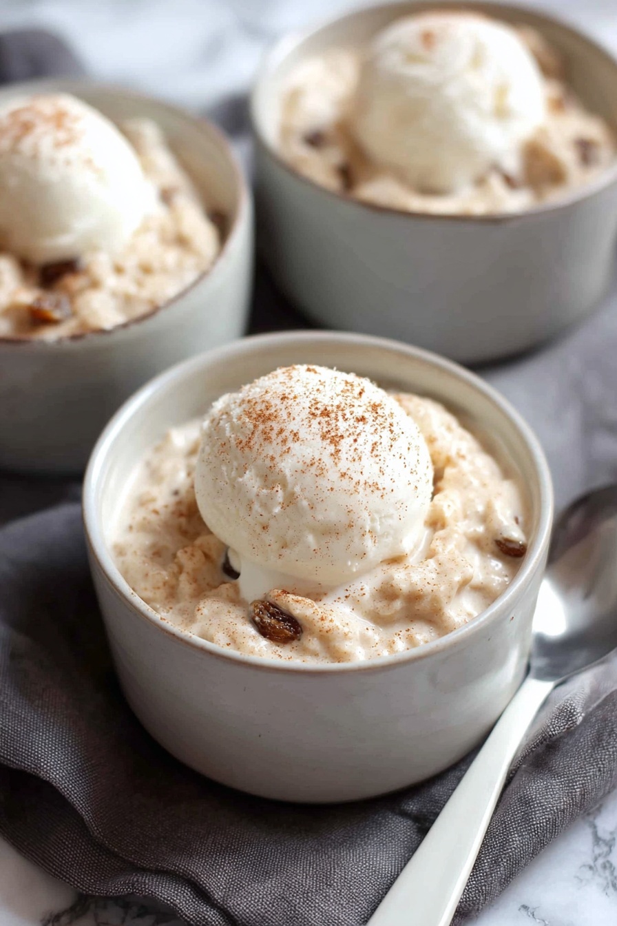 A white bowl filled with a thick creamy oatmeal mixture that has a light tan color with soft, chunky texture and dark brown raisins mixed throughout. On top of the oatmeal is a single scoop of smooth, pale white ice cream with a light dusting of brown cinnamon powder. A silver spoon is partially submerged in the oatmeal on the right side of the bowl. The bowl sits on a soft pink cloth with a white marbled surface underneath. Photo taken with an iphone --ar 2:3 --v 7 - Creamy Cinnamon Rice Pudding, cinnamon rice pudding, easy rice pudding, comforting dessert, cozy cinnamon rice pudding