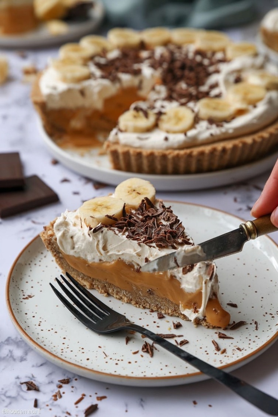A slice of pie sits on a white plate with brown speckles, showing three layers: a crunchy light brown crust, a smooth caramel-colored filling with banana slices, and a creamy white topping sprinkled with dark chocolate curls. Behind it, the rest of the pie with the same layers is on a white plate, with one slice missing. A woman’s hand holds a silver knife with some pie filling and cream on it. A black fork rests on the plate near the slice. The background is a white marbled surface with some chocolate shavings scattered around. Photo taken with an iphone --ar 2:3 --v 7 - No-Bake Banoffee Pie, Banoffee Pie without oven, Easy Banoffee Dessert, No-Bake Pie Recipes, Caramel Banana Pie