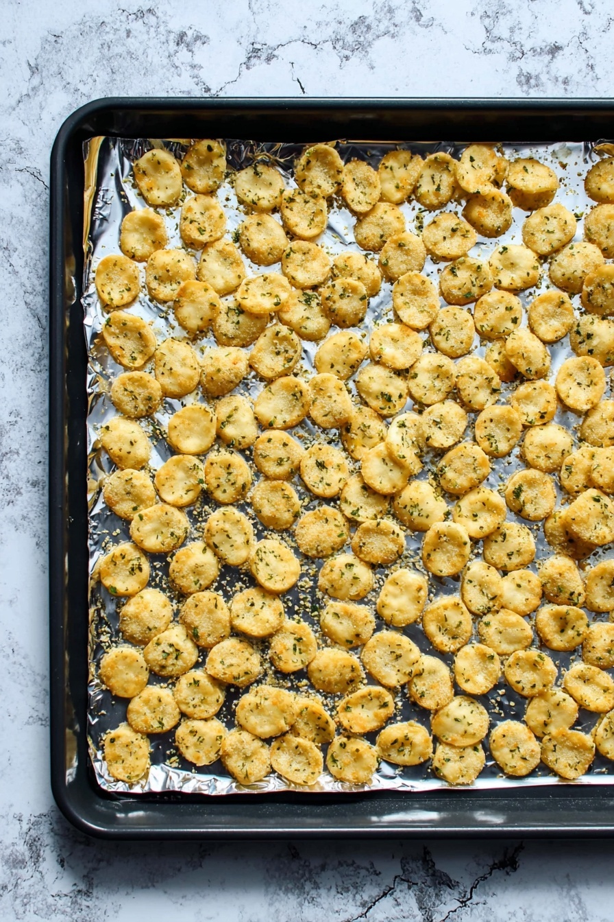 A black baking tray lined with shiny aluminum foil holds a single flat layer of small, round, golden-brown crackers sprinkled with green herbs. The crackers are spread evenly, with slight overlaps in places, showing a crispy and slightly rough texture. The background features a white marbled surface that contrasts with the baking tray. photo taken with an iphone --ar 2:3 --v 7 - Best Ranch Oyster Crackers, Ranch Oyster Crackers ingredients, how to make ranch oyster crackers, easy oyster cracker snacks, flavorful oyster crackers