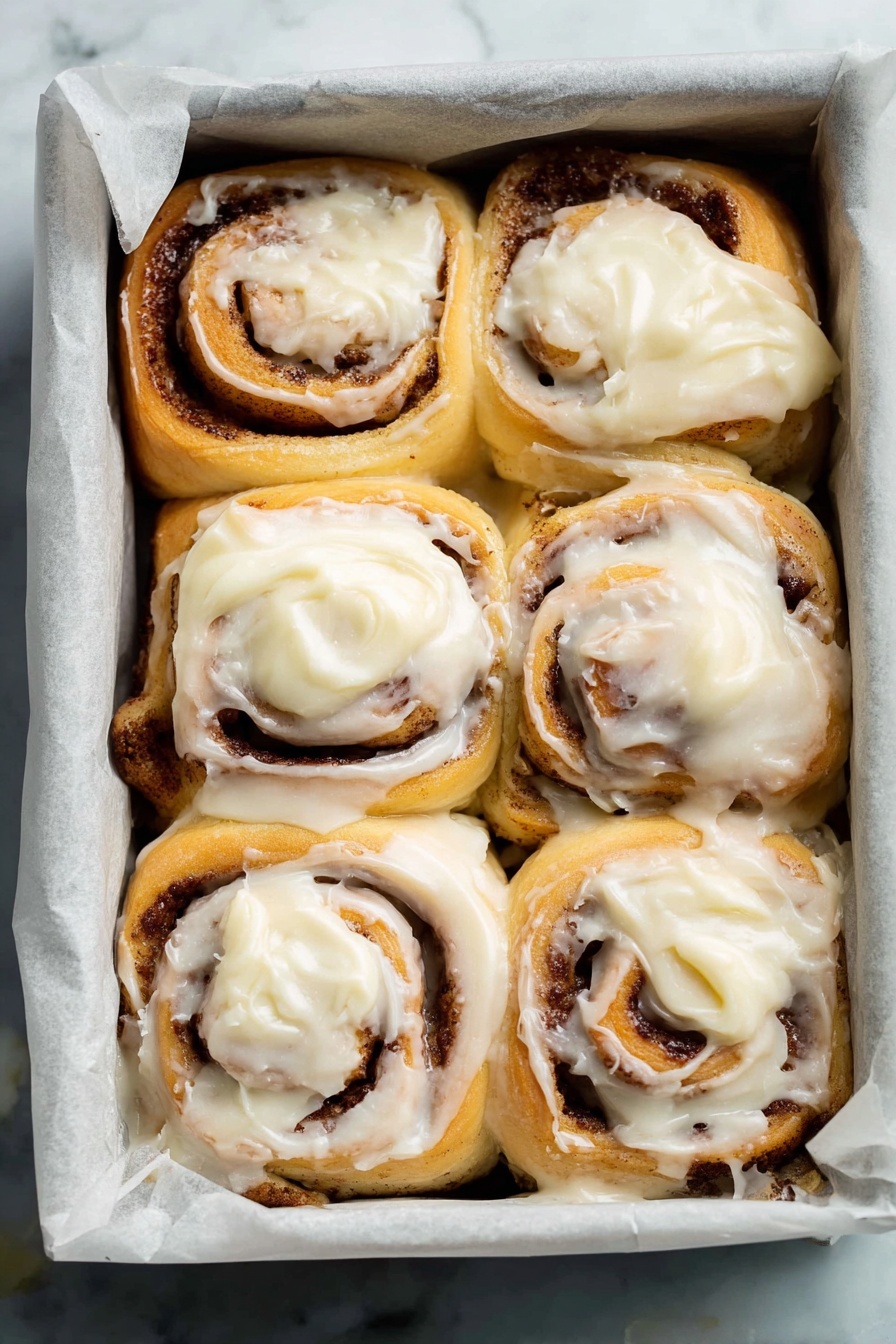 This image shows eight cinnamon rolls in a square baking pan lined with white parchment paper, placed on a white marbled surface. Each roll has two visible layers: the base layer is soft, light golden dough spiraled with a dark brown cinnamon filling, and the top layer is thick, creamy white frosting spread unevenly, melting slightly into the rolls. The rolls are packed closely together, creating a warm, inviting look with some frosting softly dripping between them. Photo taken with an iphone --ar 2:3 --v 7 - Ultimate Fluffy Cinnamon Rolls, soft cinnamon rolls, homemade cinnamon rolls, easy cinnamon roll recipe, fluffy breakfast treats