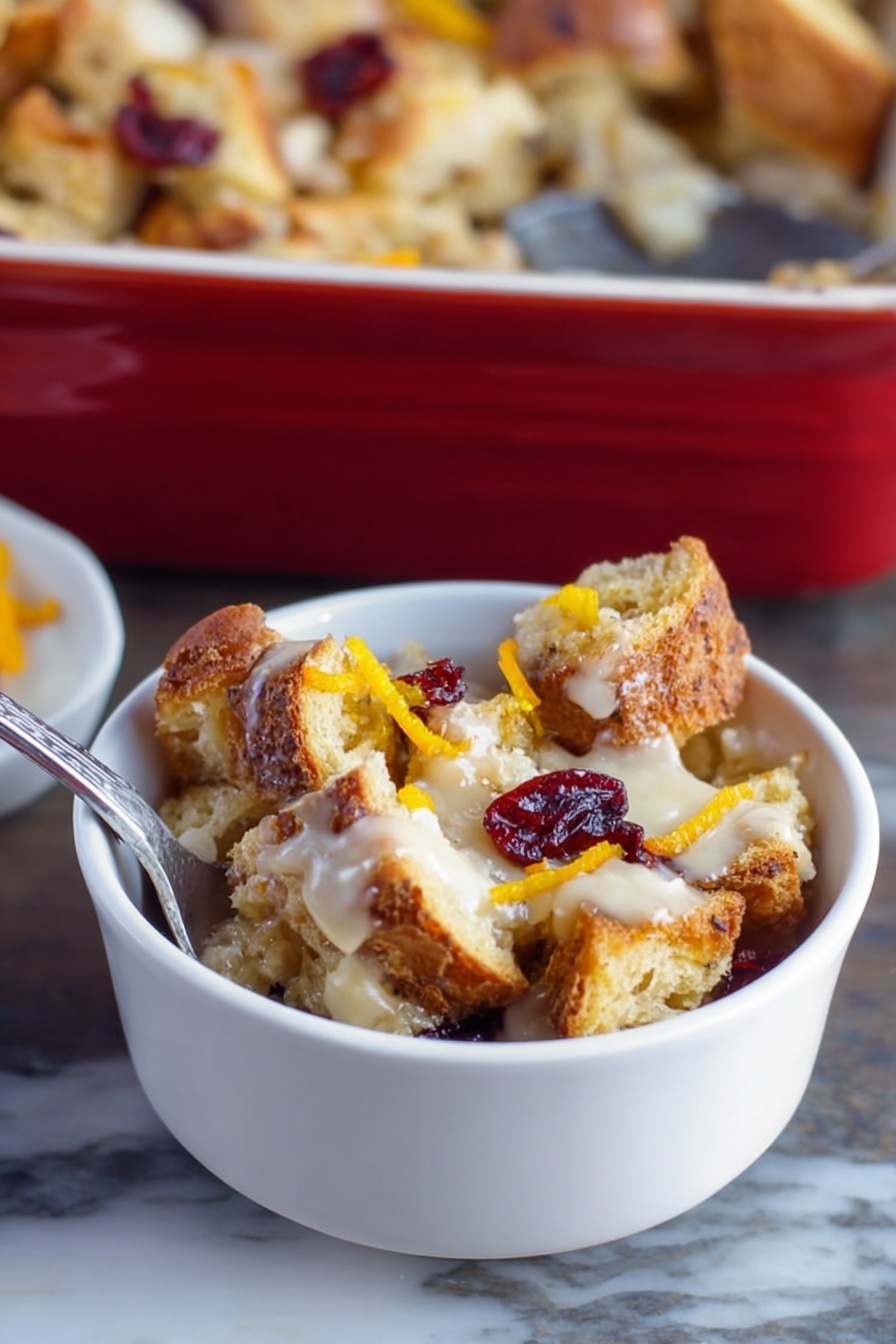 A small white bowl filled with several uneven layers of toasted bread pieces that are golden brown and crispy, mixed with a creamy, light-colored sauce spread unevenly throughout, topped with a few dried red cranberries and thin strips of bright orange zest, placed on a white marbled surface with a silver spoon inside the bowl, a larger baking dish filled with similar bread pudding in the background, photo taken with an iphone --ar 2:3 --v 7 - Cranberry Bread Pudding, holiday dessert recipes, easy bread pudding, festive cranberry dessert, cozy holiday treats