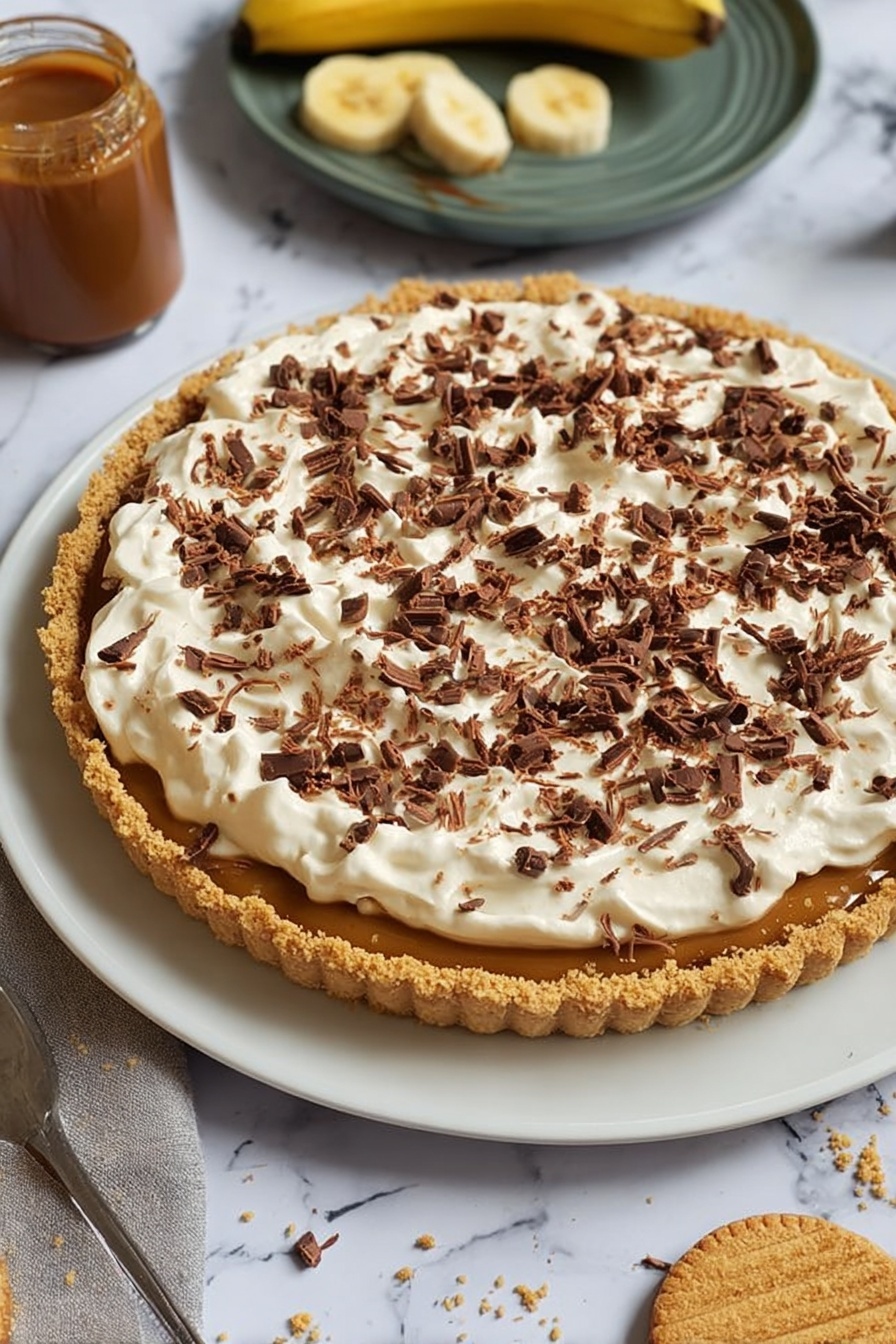 The image shows a dessert tart on a white plate with a crumbly golden brown crust forming the base, filled with a caramel-colored layer visible under a thick top layer of fluffy, white cream spread unevenly. The cream is generously covered with small, dark brown chocolate curls scattered across the surface. Around the plate, there is a jar of caramel sauce, a partially peeled banana, and a round biscuit with a bite taken out, all set on a white marbled surface. Photo taken with an iphone --ar 2:3 --v 7 - No-Bake Banoffee Pie, Banoffee Pie without oven, Easy Banoffee Dessert, No-Bake Pie Recipes, Caramel Banana Pie