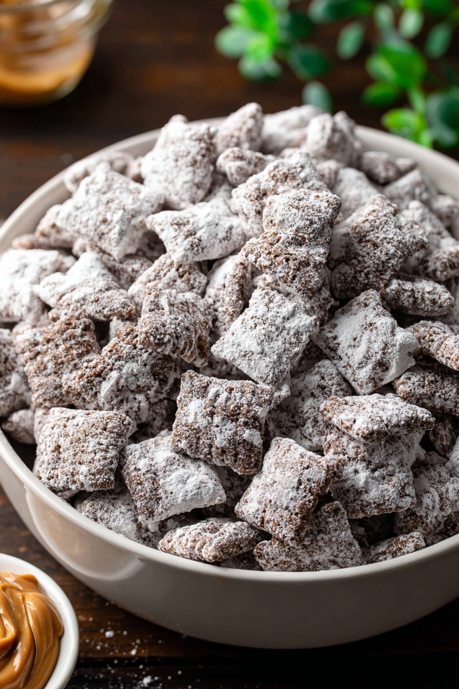 A large white bowl filled with many small square pieces of chocolate cereal covered evenly in thick white powdered sugar. The cereal pieces have a rough textured surface with some visible chocolate underneath the sugar. The bowl is placed on a dark wooden surface with a small dish of peanut butter visible in the lower left corner, and blurred green leaves in the upper left background. The image is close up showing the detailed texture of the cereal pieces. photo taken with an iphone --ar 2:3 --v 7 - Easy Chocolate Peanut Butter Puppy Chow, peanut butter puppy chow, no-bake muddy buddies, quick chocolate puppy chow, beginner-friendly snack recipes