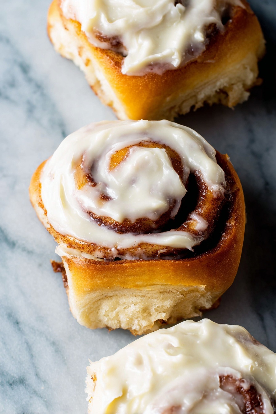 The image shows three cinnamon rolls with soft golden-brown spiral layers of dough swirled with cinnamon filling. Each roll is topped with a creamy white frosting that looks thick and smooth, spread unevenly with some parts thicker than others. The cinnamon rolls are placed on a white marbled surface that contrasts with their warm colors. The focus is close up, making the texture of the dough and frosting very clear. Photo taken with an iphone --ar 2:3 --v 7 - Ultimate Fluffy Cinnamon Rolls, soft cinnamon rolls, homemade cinnamon rolls, easy cinnamon roll recipe, fluffy breakfast treats