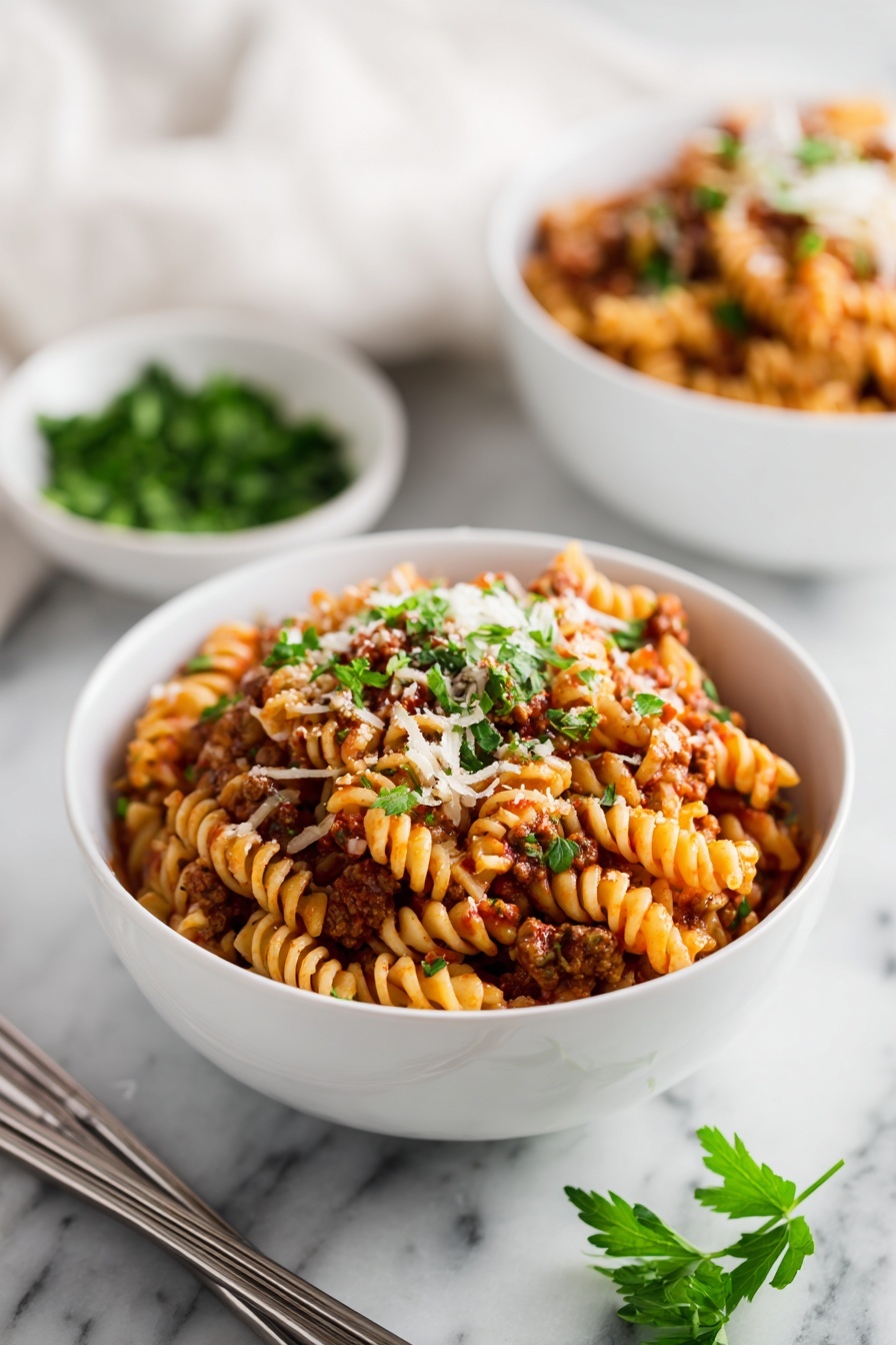 A white bowl filled with a layered pasta dish made of ruffled-edge pasta mixed with a red tomato sauce, ground meat, and small bits of melted cheese, all topped with sprinkles of chopped green parsley. Another similar bowl is in the blurred background beside a small white bowl with more parsley. The bowls are placed on a white marbled surface with a pair of silver chopsticks lying next to the front bowl. Some fresh parsley sprigs are also visible at the bottom right corner. Photo taken with an iphone --ar 2:3 --v 7 - Instant Pot Lasagna, easy Instant Pot lasagna, quick lasagna recipe, hearty Instant Pot dinner, one-pot lasagna