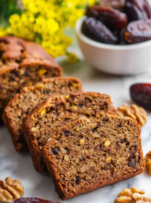 A close-up of four slices of brown nut bread lined up diagonally on a white marbled surface. Each slice shows a dense texture with visible dark brown nuts and darker bits inside the bread. Around the slices are loose whole walnuts and dark brown dried dates scattered. In the background, there is a white bowl filled with more dried dates and a soft-focused cluster of yellow flowers. The scene is bright and natural. photo taken with an iphone --ar 2:3 --v 7 - Date Nut Bread, moist date nut bread, homemade date bread, healthy date nut loaf, easy date bread recipe