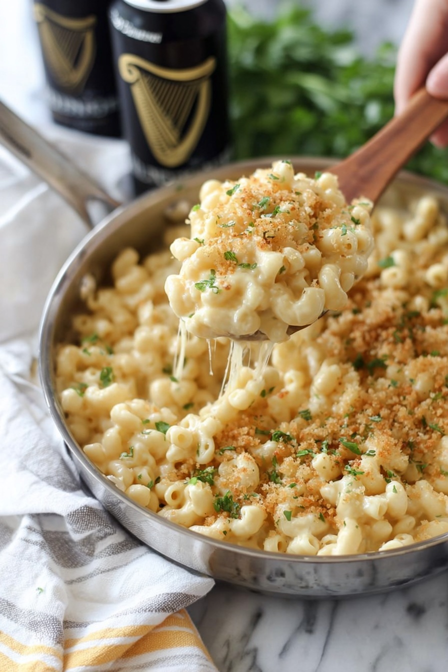 A close-up view of a shiny silver pan filled with creamy macaroni and cheese, with small pasta curls covered in a smooth, light yellow cheese sauce. The top has a sprinkle of golden brown breadcrumbs and bits of green herbs, adding texture. A wooden spoon, held by a woman's hand, lifts a portion of the macaroni, showing stretchy cheese strings between the pan and the spoon. In the background, some green leaves and two black cans with gold writing are slightly out of focus. The whole scene is set on a white marbled surface with a white cloth that has gray and yellow stripes. photo taken with an iphone --ar 2:3 --v 7 - Guinness Irish Cheddar Mac and Cheese, Irish cheddar mac and cheese, Guinness beer infused mac and cheese, classic Irish cheese pasta, creamy Guinness mac and cheese