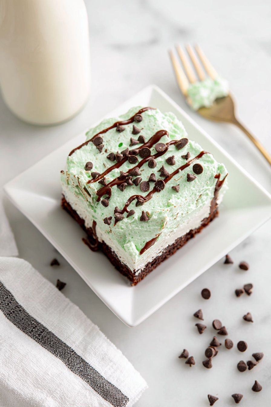 The image shows a square piece of cake on a white square plate, with three visible layers: a bottom moist dark brown cake layer, a middle light brown layer, and a thick top layer of light mint green cream frosting. The frosting is textured and decorated with scattered small dark brown chocolate chips and thin drizzles of shiny melted chocolate across the surface. A fork with some green frosting remains lies nearby on the white marbled surface, around which a few chocolate chips are also scattered. A bottle with a light creamy-colored drink is visible in the top-left corner, and a white towel with black stripes is placed at the bottom. Photo taken with an iphone --ar 2:3 --v 7 - Mint Chocolate Ice Cream Sandwich Cake, easy ice cream cake recipes, no-bake summer desserts, mint chocolate dessert ideas, crowd-pleasing ice cream cakes