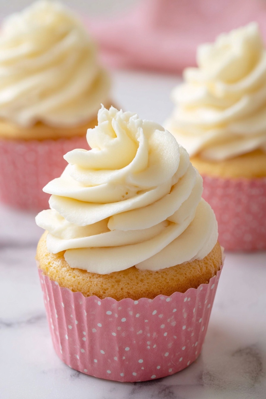 The image shows a close-up of a cupcake with three layers visible. The bottom layer is a light golden cake wrapped in a pink cupcake liner with small white polka dots. The top layer is a thick swirl of creamy white frosting, smoothly textured with soft peaks rising to a point. Behind it, two more cupcakes of the same kind are slightly out of focus, sitting on a white marbled surface. The lighting is soft, highlighting the frosting’s smoothness and the cake’s light texture. photo taken with an iphone --ar 2:3 --v 7 - Moist Vanilla Cupcakes, fluffy vanilla cupcakes, easy vanilla cupcake recipe, tender vanilla cupcakes, vanilla cupcake baking