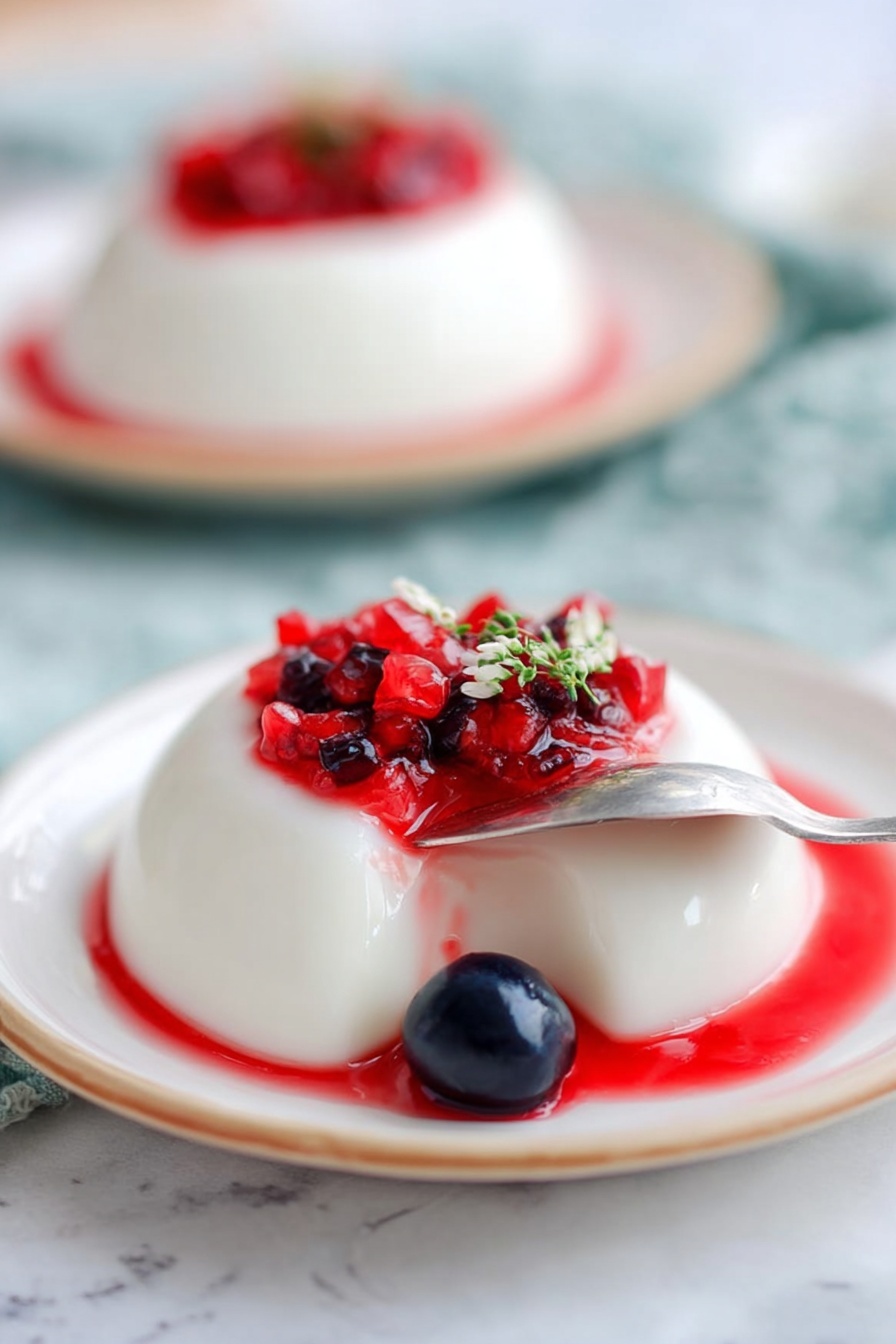 The image shows a smooth, white panna cotta dessert with one bite taken out from the front side, revealing its creamy texture. On top, there is a layer of small cubed bright red strawberries and a few whole shiny dark blue blueberries, along with a small green sprig of herb with tiny purple flowers as garnish. The panna cotta sits on a white plate with a green center and a thin golden rim. The plate is on a white marbled surface with soft, blurred pastel colors in the background. Photo taken with an iphone --ar 2:3 --v 7 - Greek Yogurt Panna Cotta with Thyme Berries, Greek Yogurt Panna Cotta, Healthy Italian Desserts, Easy Summer Dessert Recipes, Elegant Berry Panna Cotta
