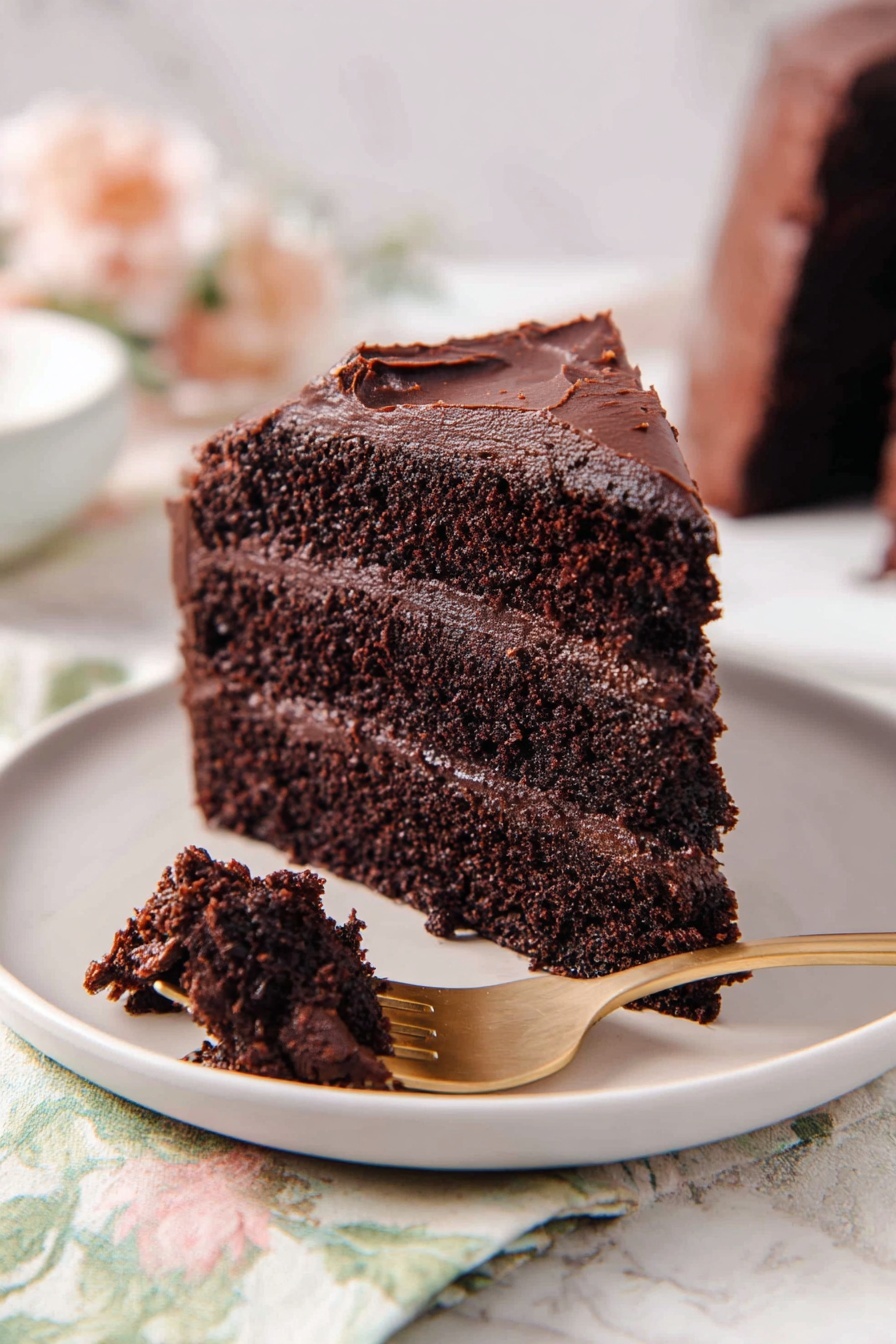 A moist chocolate cake slice with three thick layers, each dark brown and rich in texture, separated by smooth and creamy chocolate frosting layers. The outside is covered with the same dark chocolate frosting, slightly uneven but thickly spread. A small piece of the cake is held on a gold fork in the front, showing the crumbly, soft texture inside. The cake sits on a white plate, placed on a white marbled surface with a soft floral cloth nearby. photo taken with an iphone --ar 2:3 --v 7 - Decadent Chocolate Cake, Chocolate Cake Recipe, Moist Chocolate Cake, Rich Chocolate Cake, Easy Chocolate Cake