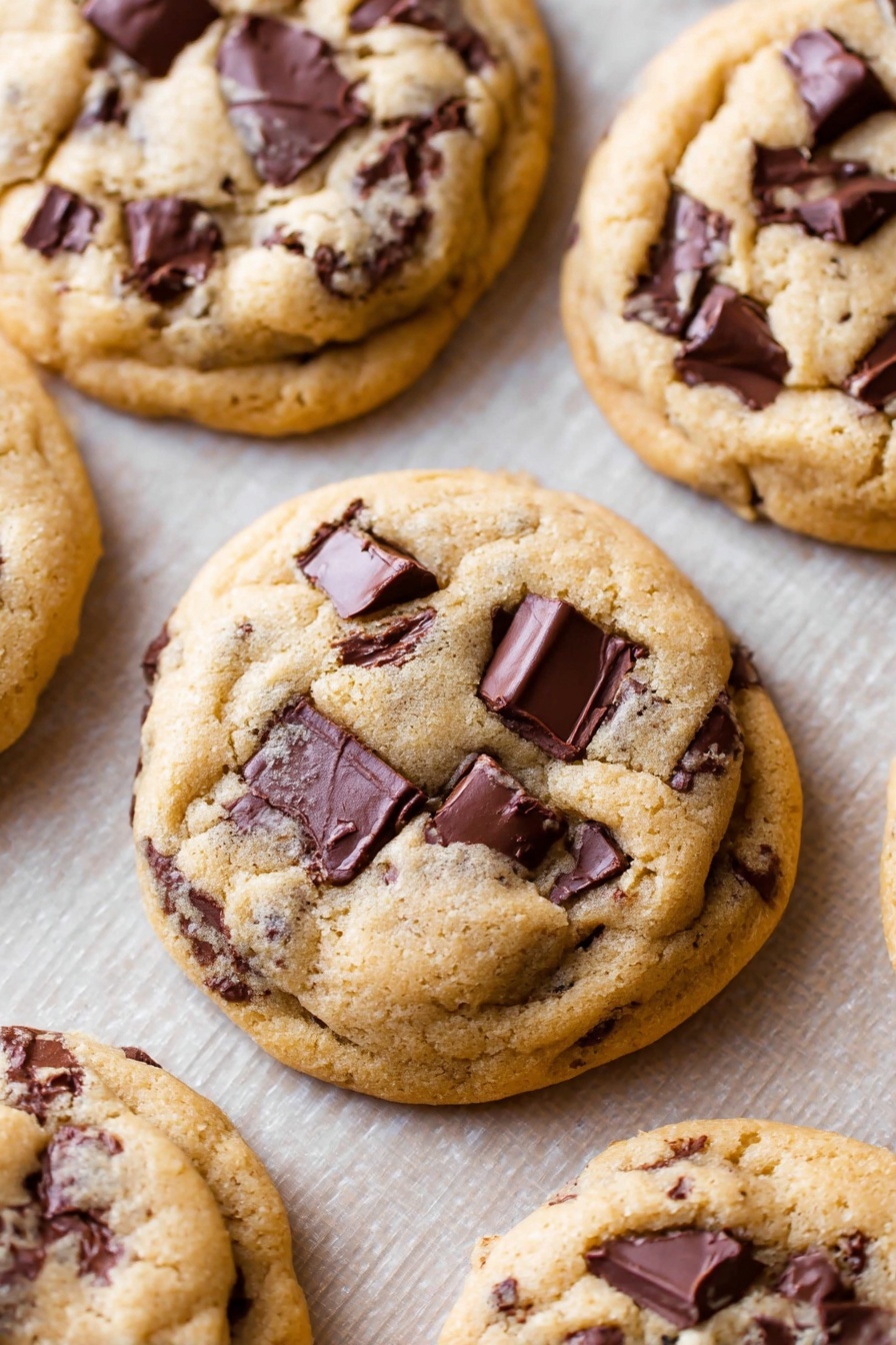 The image shows a close-up of soft, round chocolate chip cookies arranged on a baking sheet. Each cookie has a light golden-brown dough base with large, uneven chunks of dark chocolate embedded throughout, creating a textured look. The dough looks slightly raised and smooth with some small cracks and crevices. The cookies are stacked closely, with the front cookie prominently showing the rich chocolate pieces on top and within the dough. The baking sheet underneath has a grid-like pattern. The background is a white marbled texture. Photo taken with an iphone --ar 2:3 --v 7 - Chewy Chocolate Chip Cookies, easy chocolate chip cookie recipe, how to make chewy cookies, soft chocolate chip cookies, homemade cookie recipe