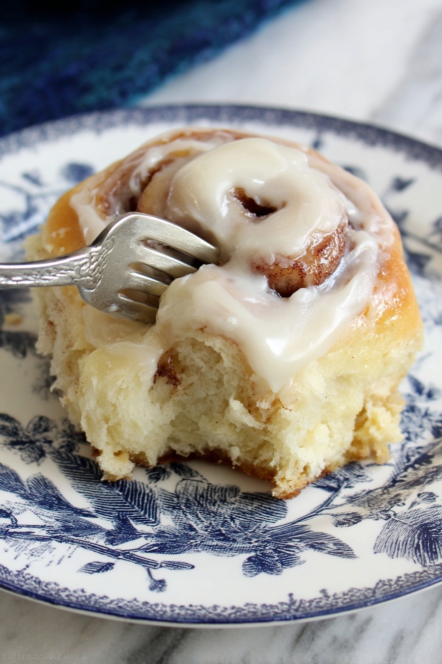 A soft cinnamon roll with three visible layers sits on a white plate with a blue floral design. The bottom layer is a golden brown dough that looks fluffy and light. The middle layer shows a swirl of cinnamon filling with a slightly darker brown color. The top layer is covered in a thick, creamy white icing that shines and drapes over the roll's edges. A silver fork presses gently into the roll’s side, pulling it slightly apart to reveal the soft texture inside. The plate is placed on a white marbled surface. Photo taken with an iphone --ar 2:3 --v 7 - Best Easy Cinnamon Rolls with Cream Cheese Icing, easy cinnamon roll recipe, homemade cinnamon rolls, quick cinnamon roll recipe, fluffy cinnamon rolls with frosting