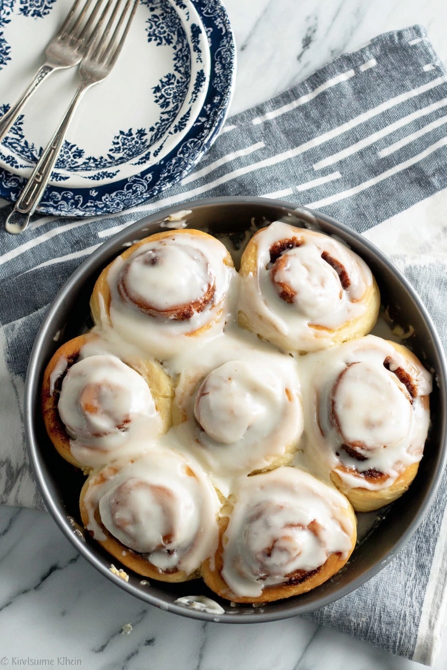 A round metal pan holds eight soft cinnamon rolls arranged in a circle, each covered with a thick layer of white icing that softly drips down the sides. The rolls are light golden brown with visible swirls of cinnamon peeking through under the creamy frosting. The pan sits on a white marbled surface with a grey and white striped cloth underneath it. Next to the pan is a white plate with a dark blue and white patterned border, accompanied by a silver fork resting on the plate. photo taken with an iphone --ar 2:3 --v 7 - Best Easy Cinnamon Rolls with Cream Cheese Icing, easy cinnamon roll recipe, homemade cinnamon rolls, quick cinnamon roll recipe, fluffy cinnamon rolls with frosting